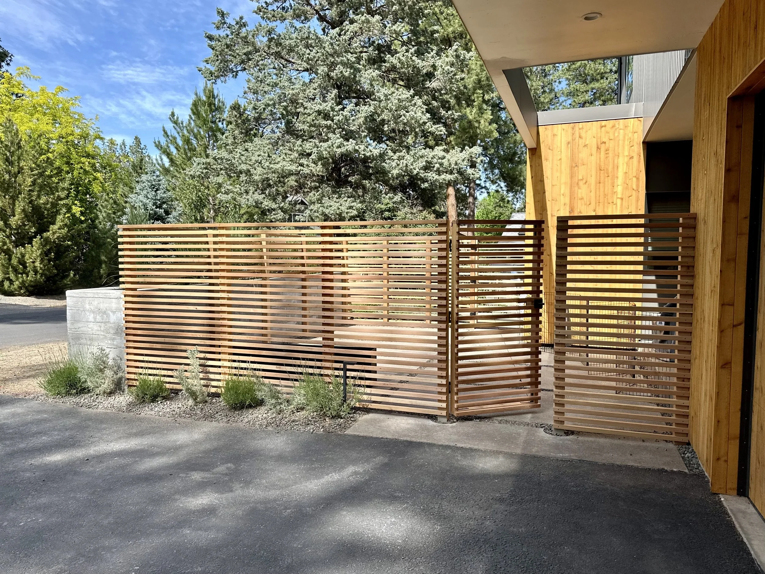 Modern residential building with wooden exterior walls and a wooden slatted privacy fence in front. The area features a paved driveway, some small green plants, and tall trees in the background under a partly cloudy sky.