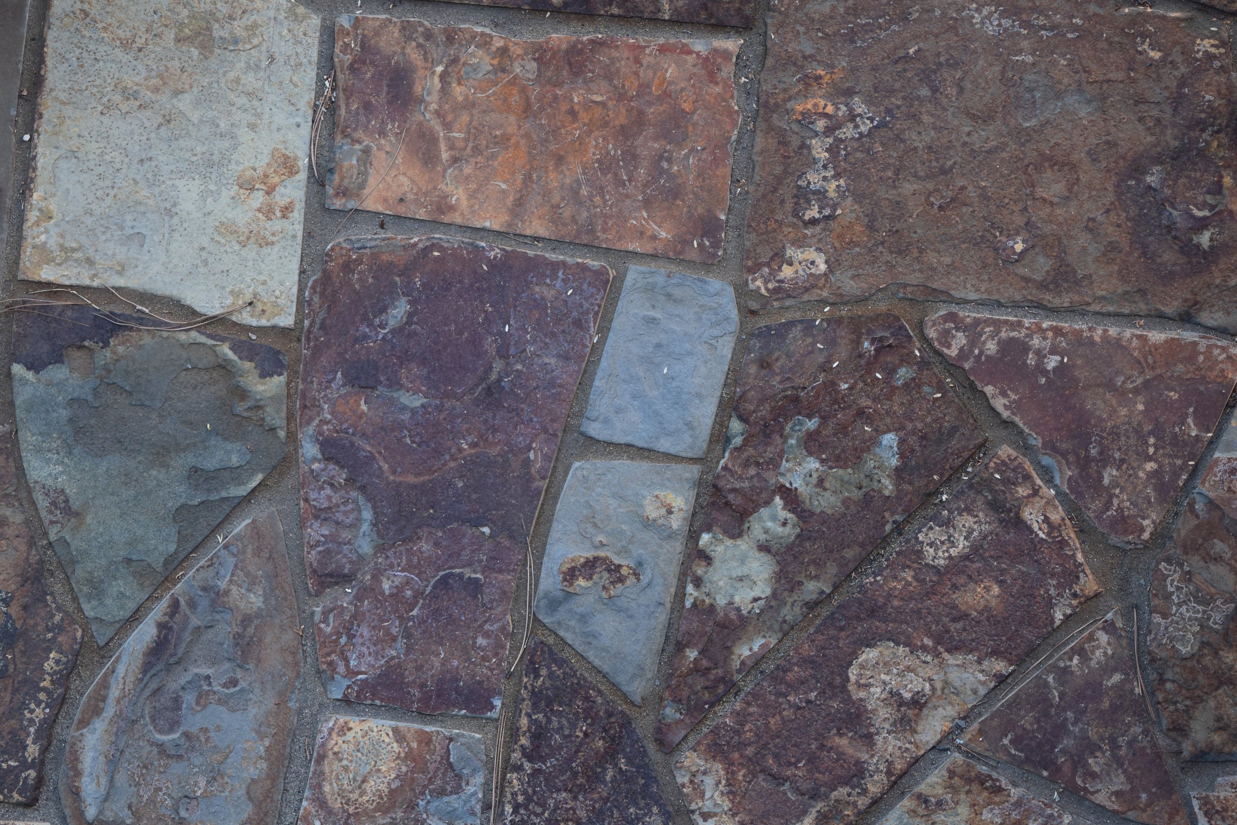 Close-up of a stone patio with irregularly shaped multicolored stones, including shades of brown, blue, gray, and reddish tones, set in grout.