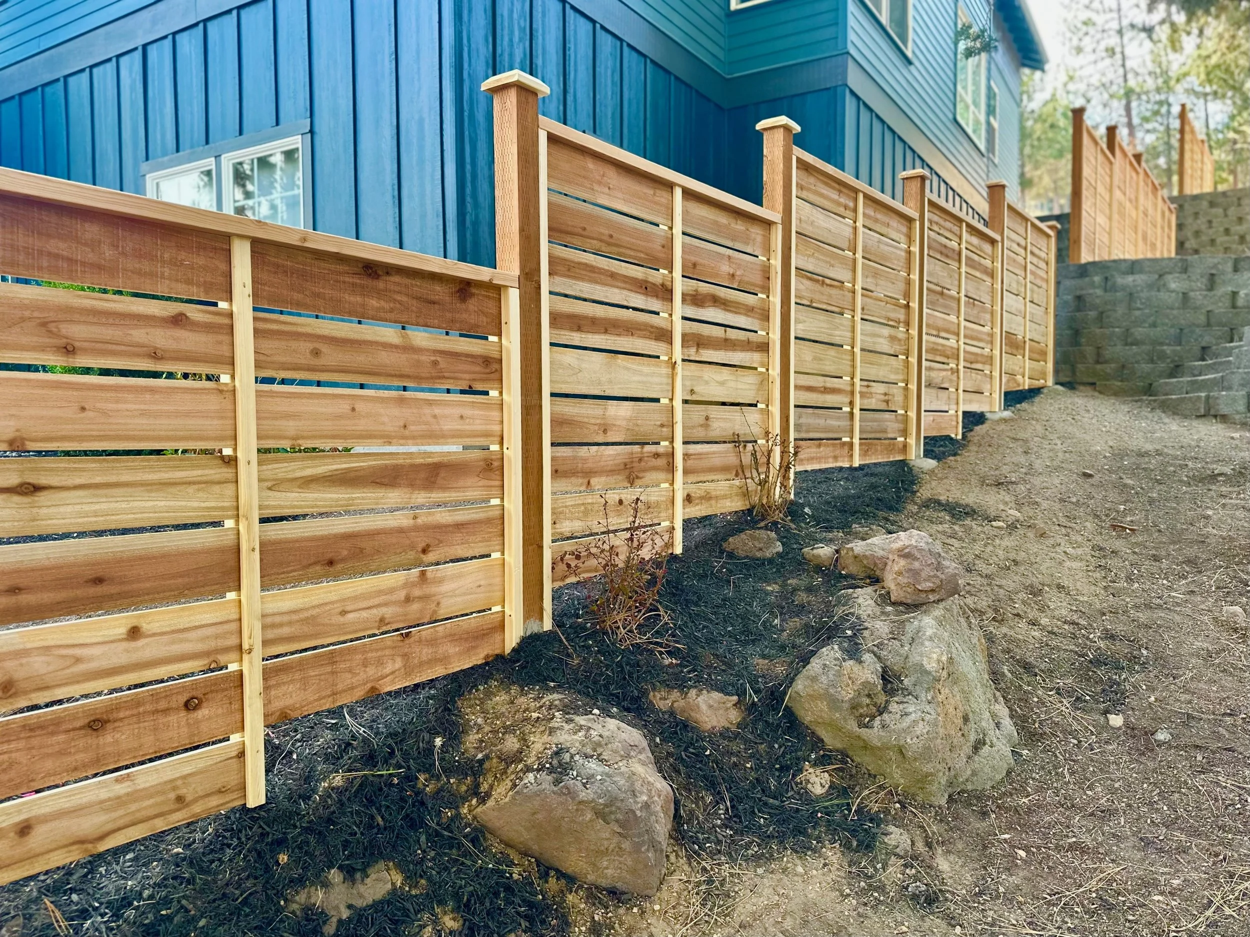 Wooden privacy fence on a sloped yard with large rocks, adjacent to a blue house with white trim, situated in a wooded area.