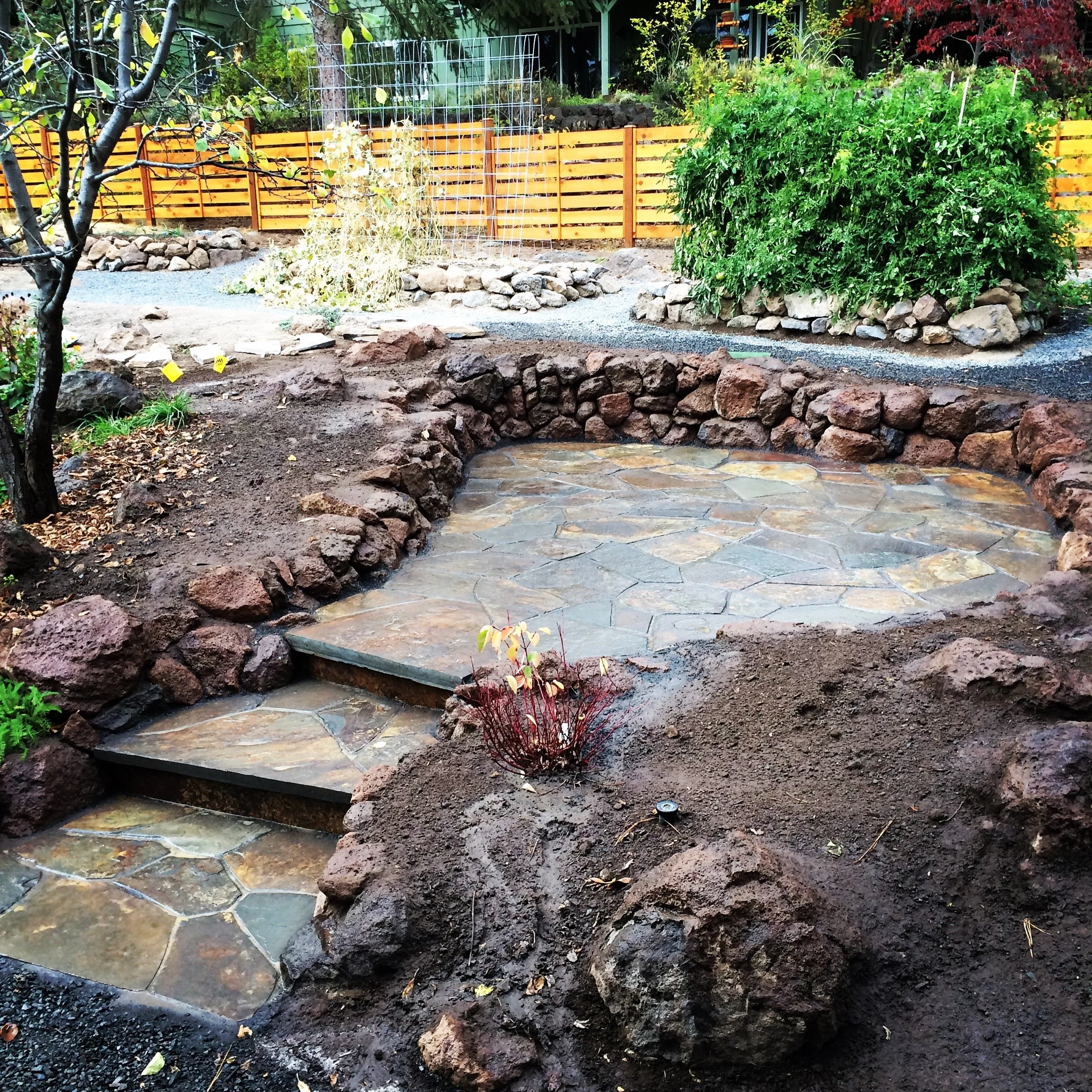 A landscaped backyard with a stone patio area surrounded by large rocks and soil. In the background, there are plants, trees, a wooden fence, and garden beds. The patio features irregularly shaped stones and a small set of steps leading to another se