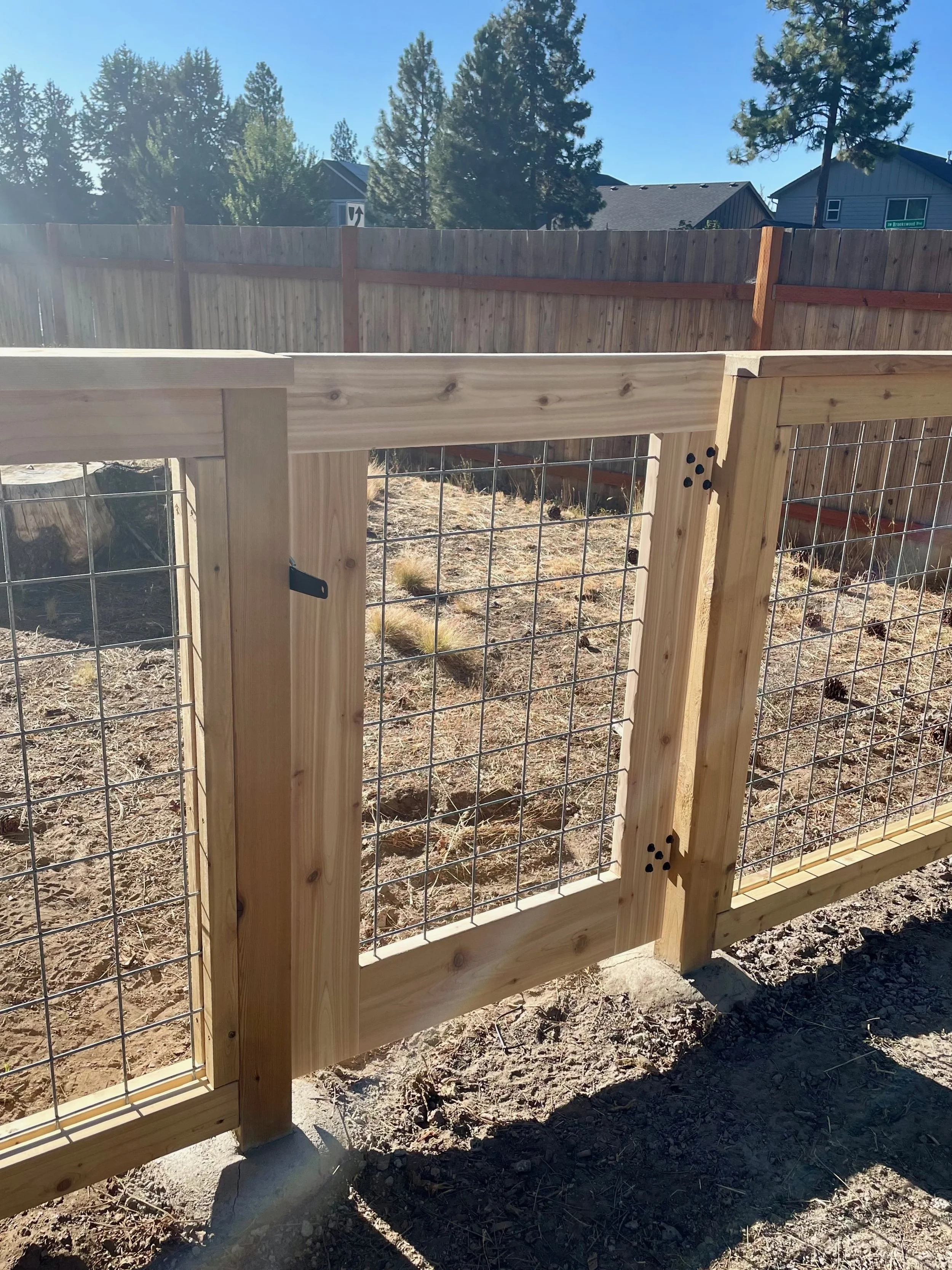 Newly built wooden fence with wire mesh panels in a backyard, under clear blue sky, with trees and houses in the background.