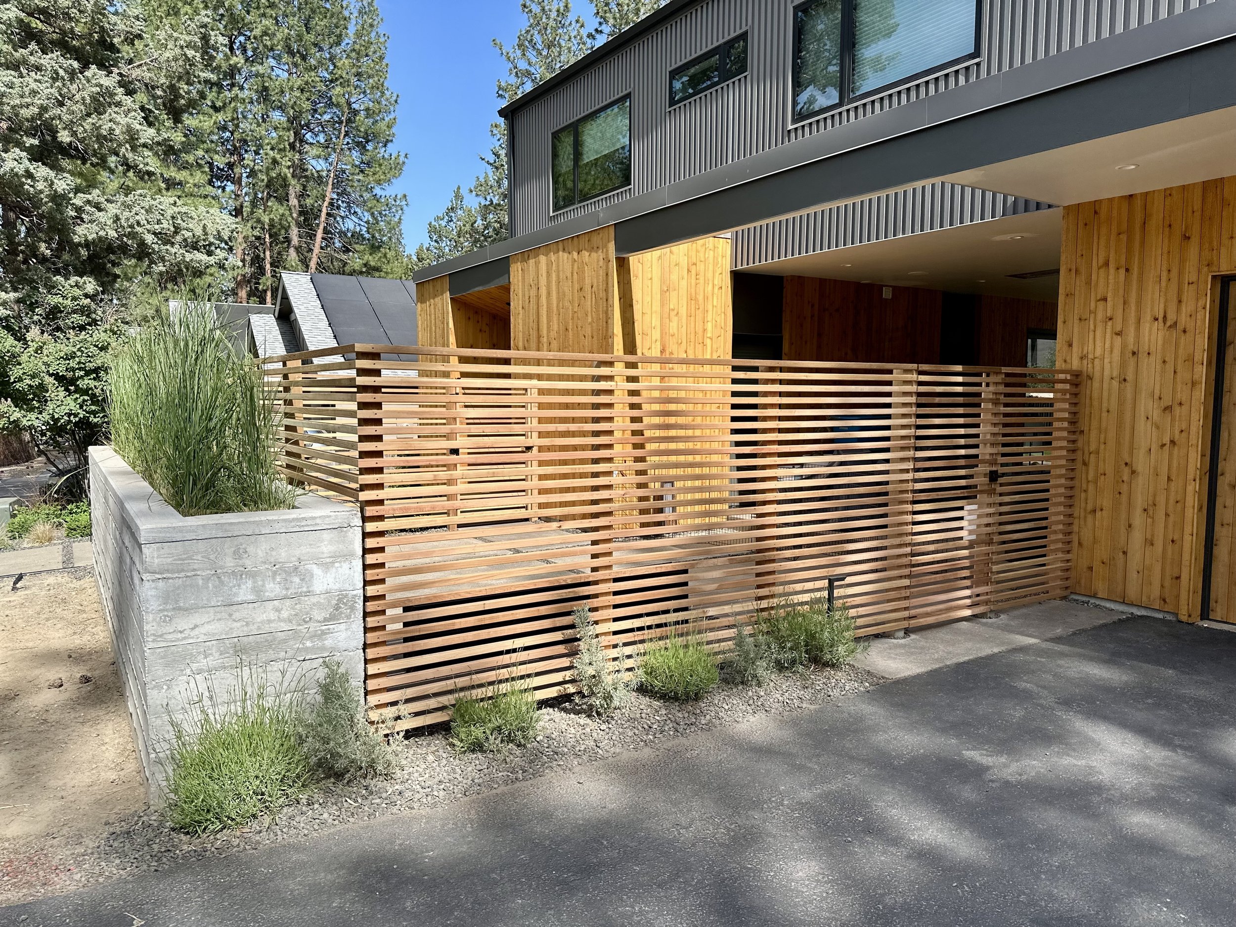 Modern building with a wooden and metal exterior, featuring a slatted wooden privacy fence and green plants in a concrete planter.