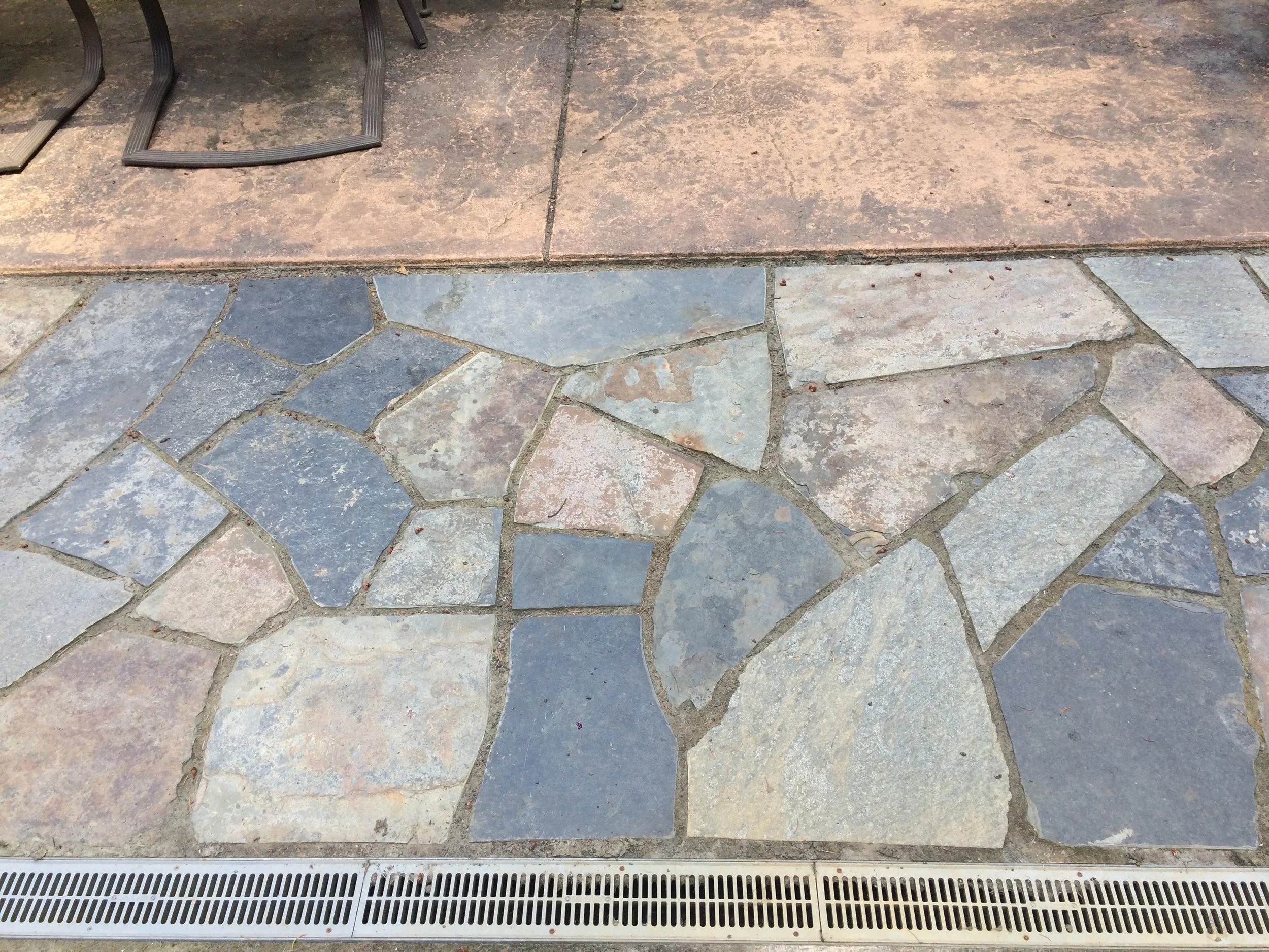 Close-up of multicolored irregular stone tiles on a patio or walkway, with a metal drain at the bottom and a section of reddish-brown paving stones at the top.