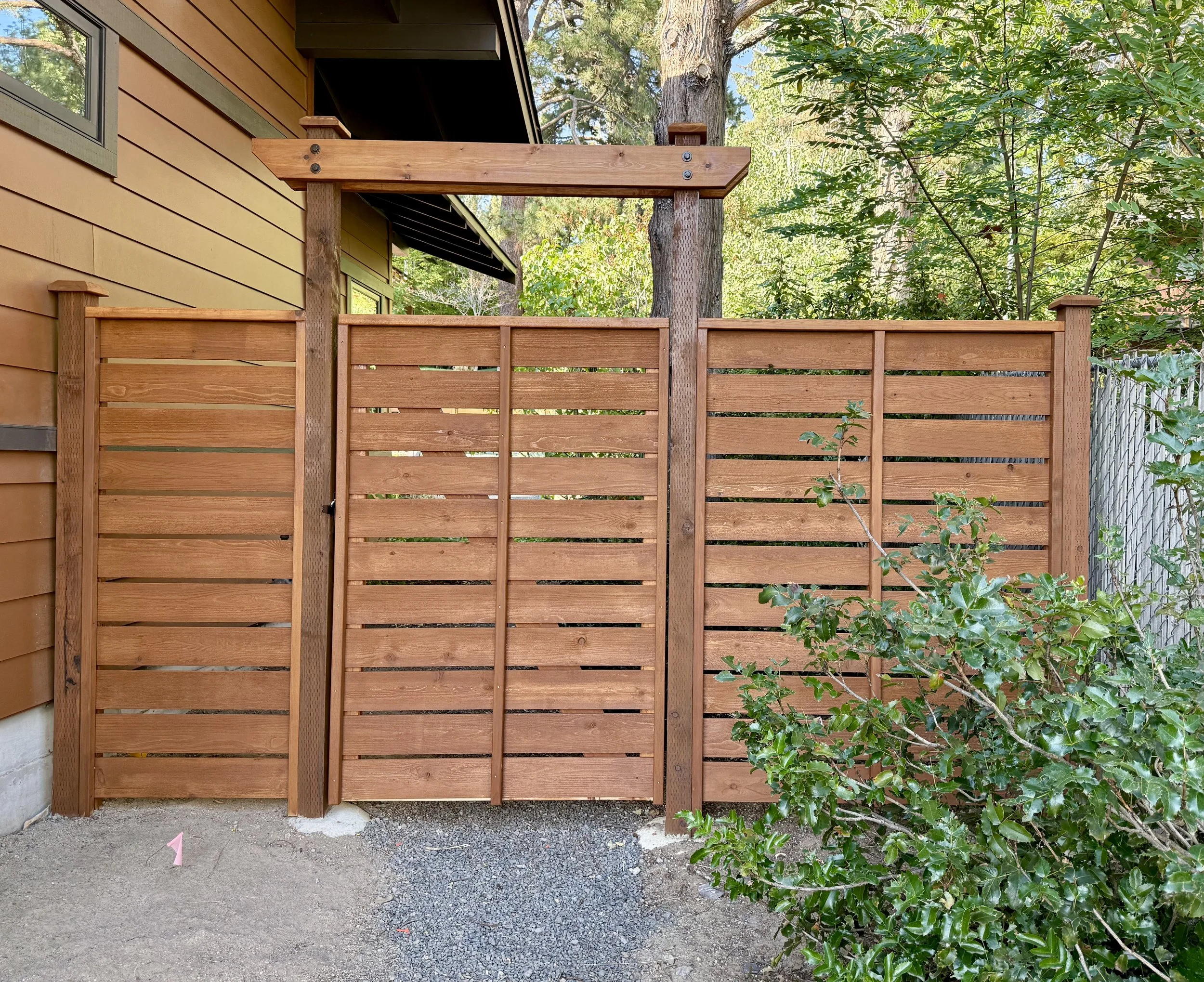 Wooden privacy fence next to a house, with a bush and trees in the background.