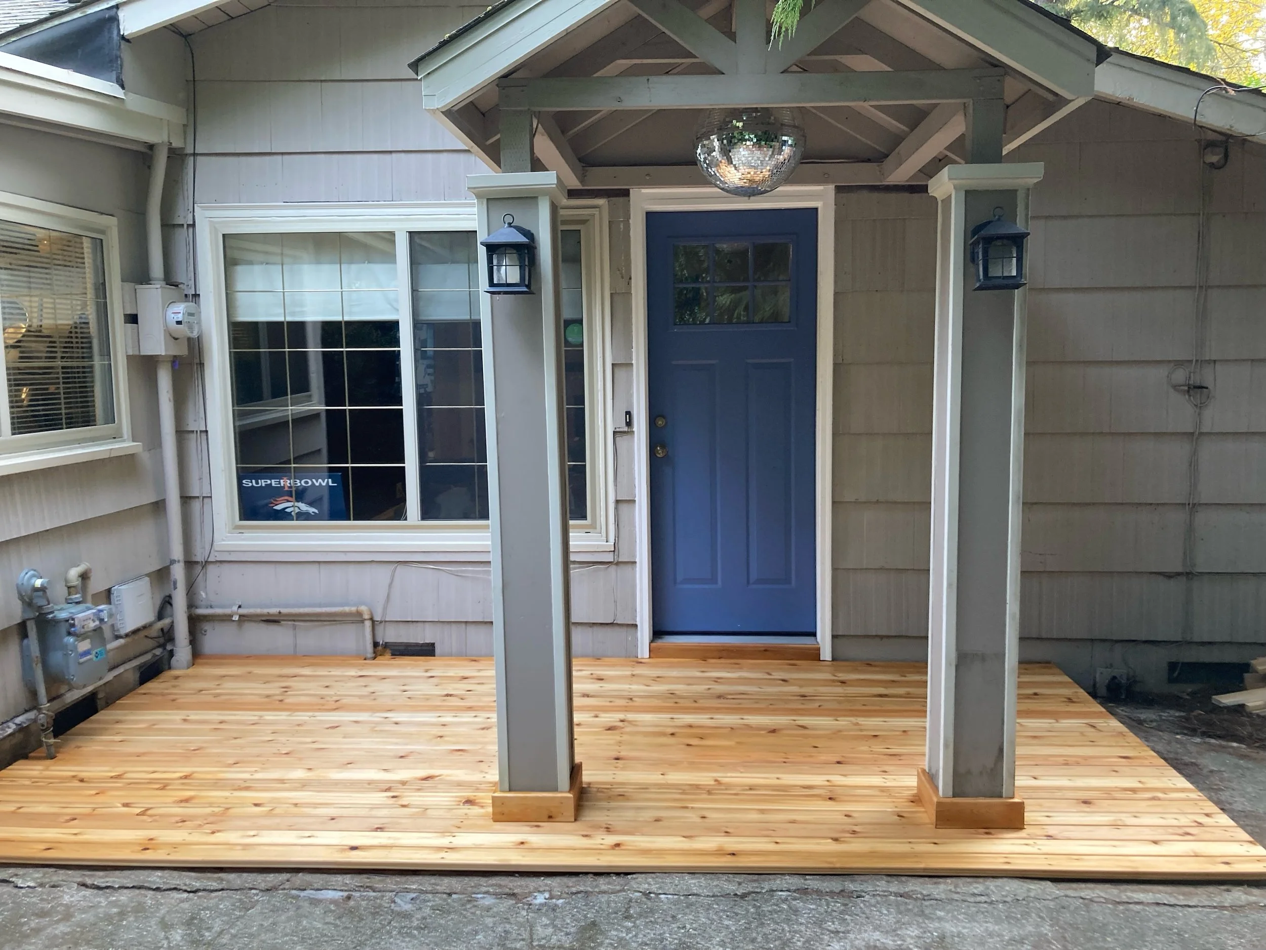 New wooden porch with a blue door, two lantern-style lights, and a small roof over the porch supported by columns, attached to a house with beige siding and a large window.