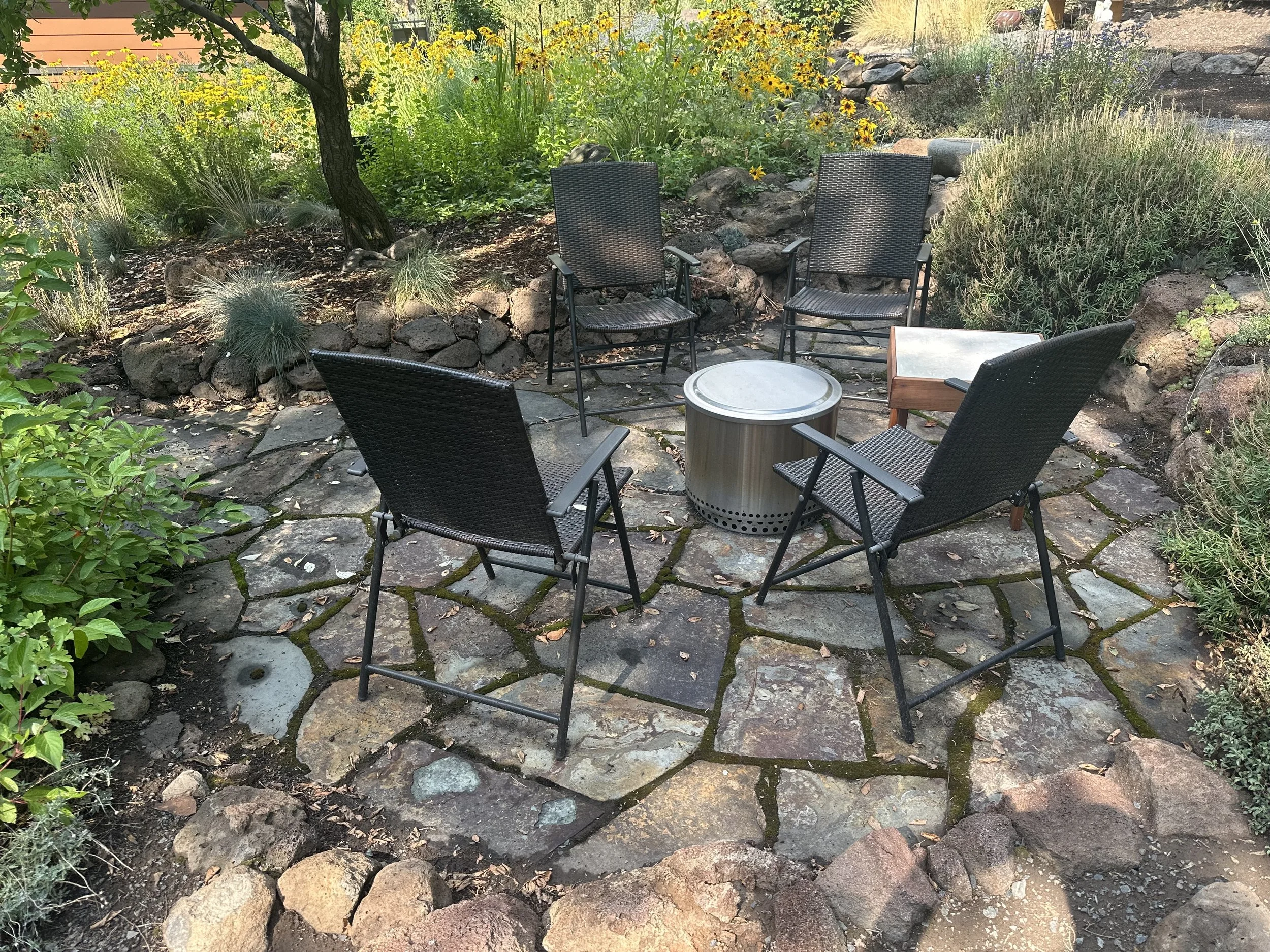 Six black outdoor chairs arranged in a circle around a round metal fire pit on a stone patio in a garden with various plants and flowers.