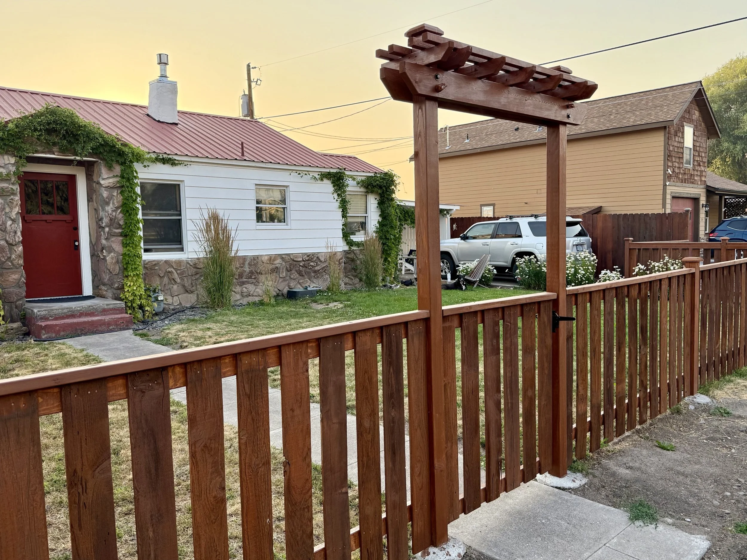 View of a backyard with a wooden fence and gate, white house with stone accents and red front door, lawn with ornamental grasses, parked white SUV, neighboring house in the background, and a sunset sky.