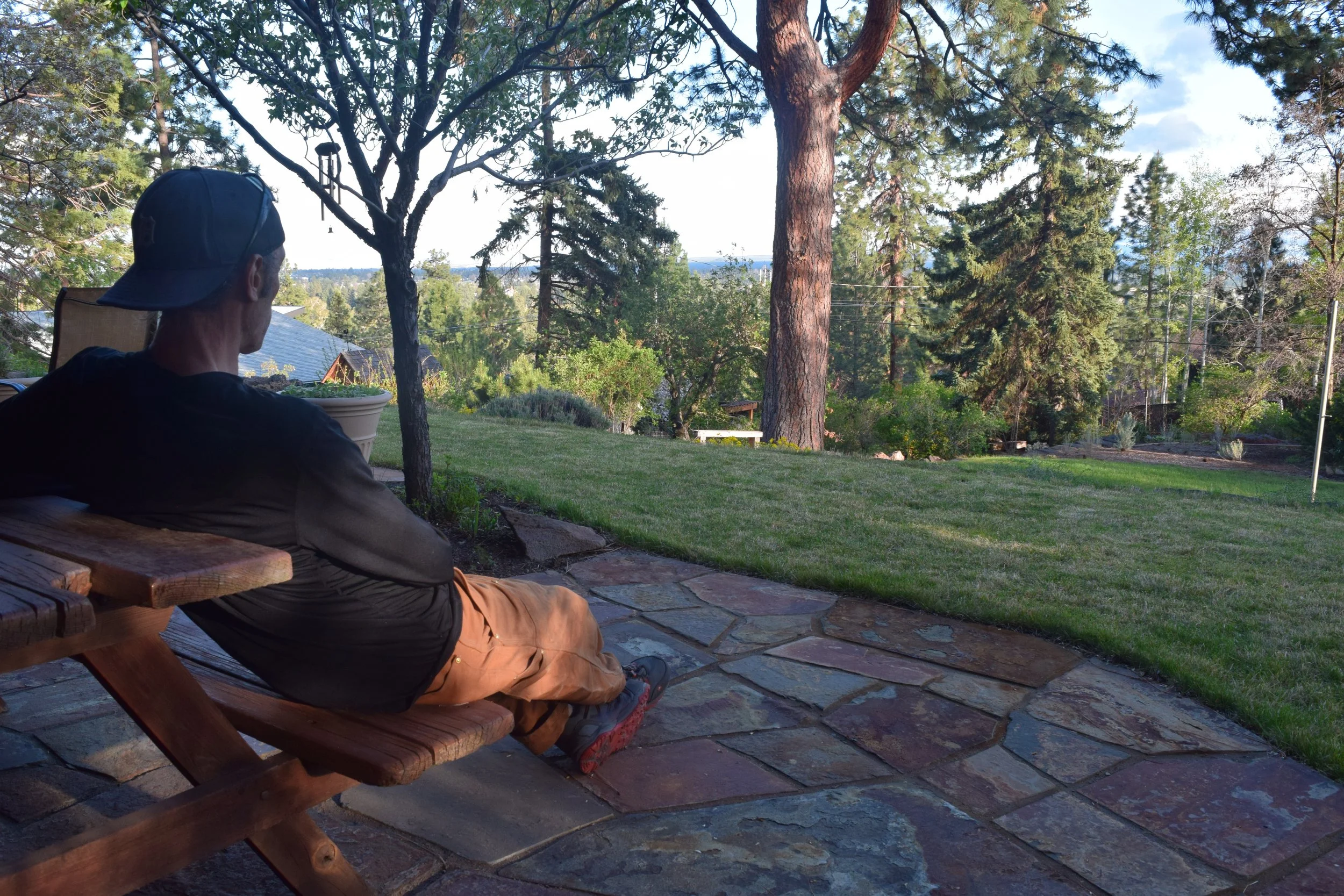 A person sitting on a wooden bench on a stone patio, overlooking a backyard with trees and a view of distant houses and hills.