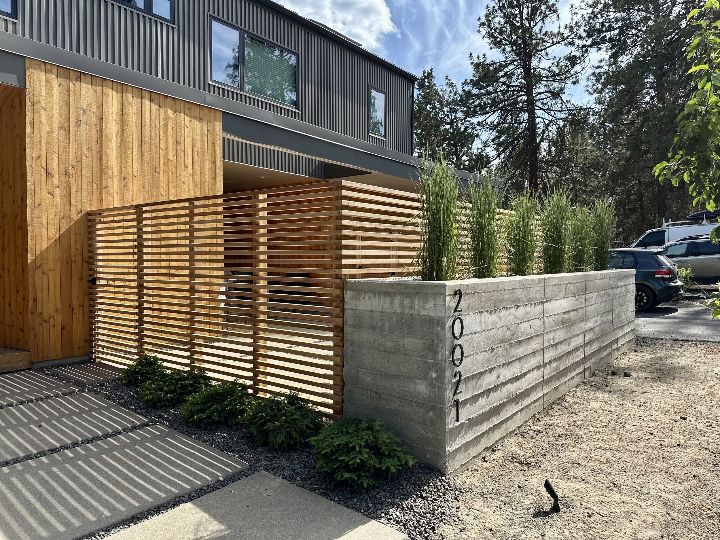 Modern building with wood and gray siding, concrete planter with tall grasses, wooden privacy slat fence, parked cars, trees, and a partly cloudy sky.