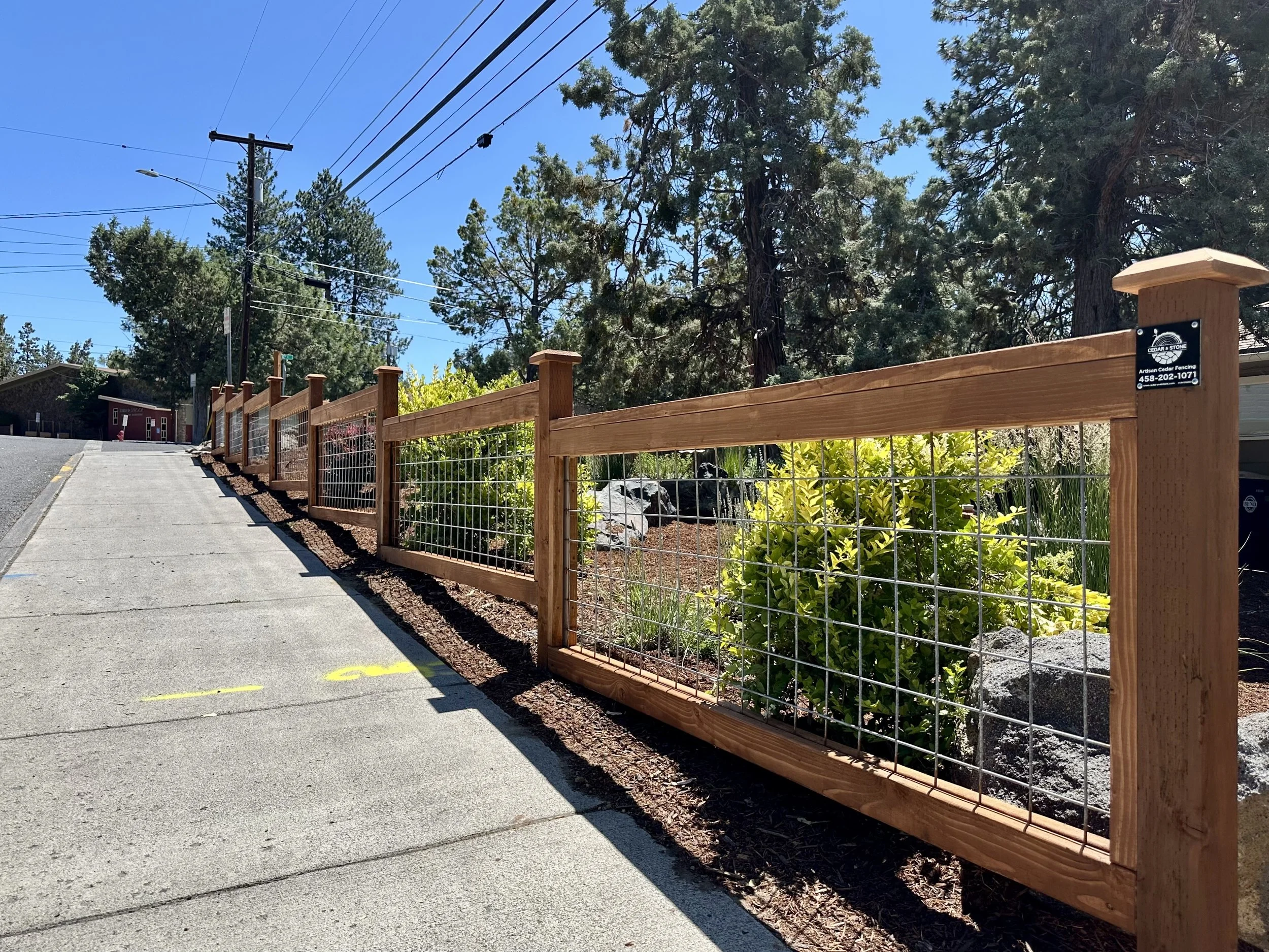 Wooden and wire fence alongside a sidewalk with trees and a small building in the background under a clear blue sky.