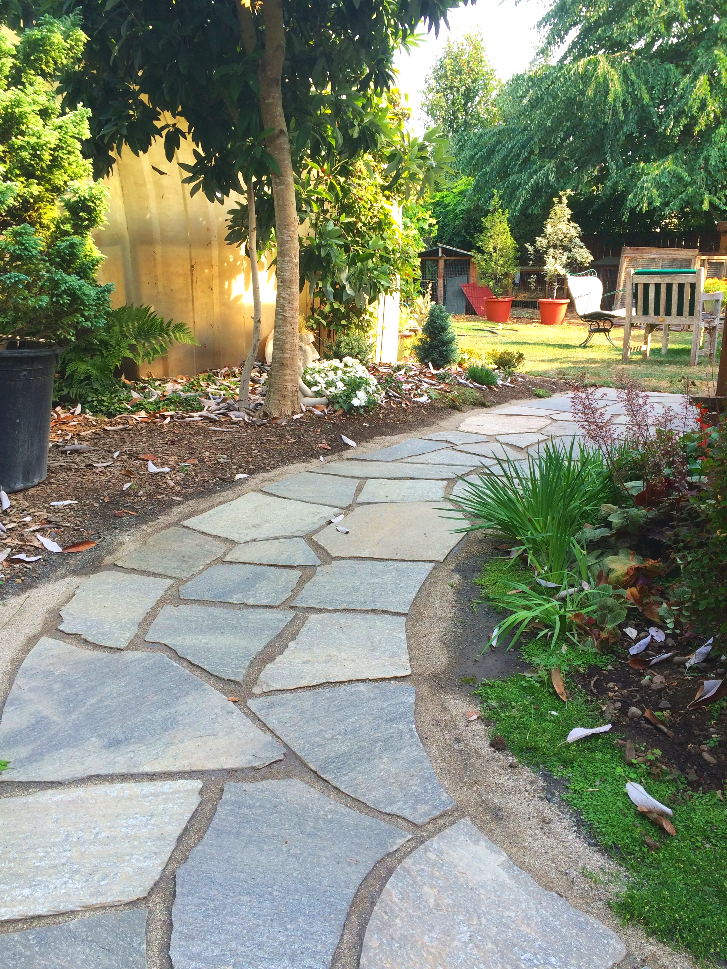 A garden pathway made of irregularly shaped gray stone pavers winding through a lush, green garden with trees, plants, and outdoor furniture.