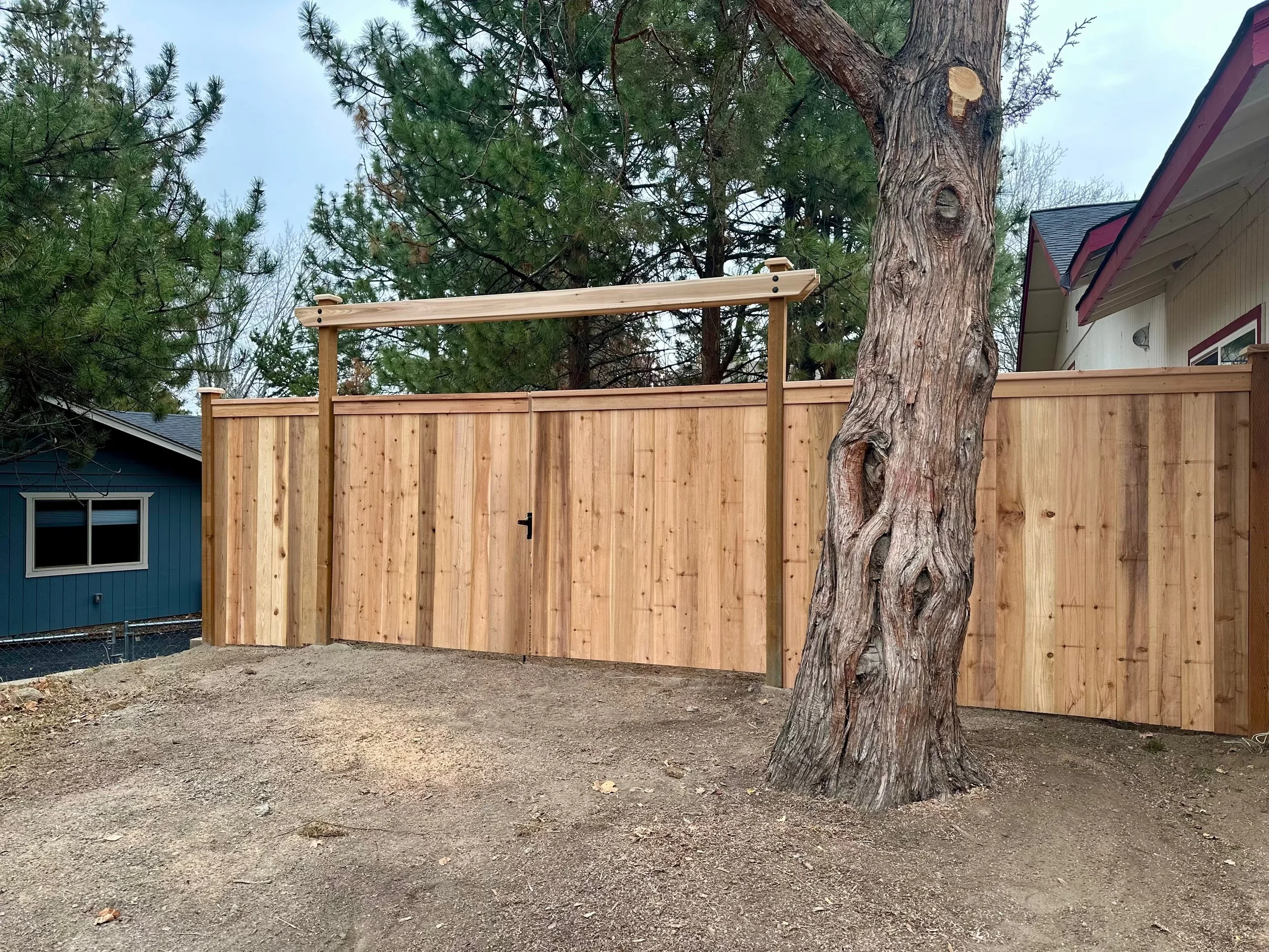 A newly built wooden fence with a gate, next to a large, partially visible tree, with a house in the background and neighboring houses partially visible on both sides.