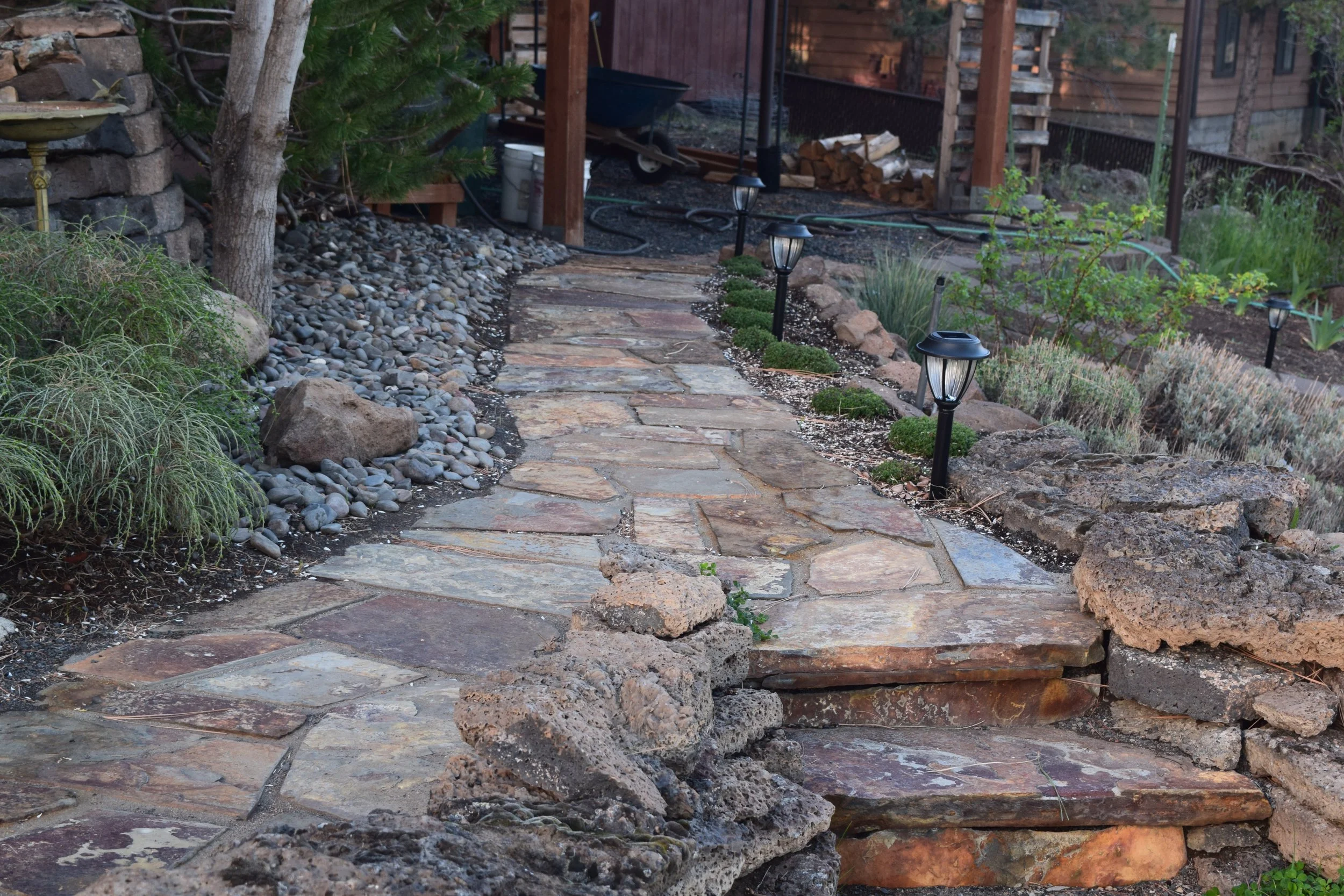A stone walkway in a backyard with garden lights along the pathway, surrounded by plants and rocks, leading to a building with a wooden deck and firewood stacked underneath.