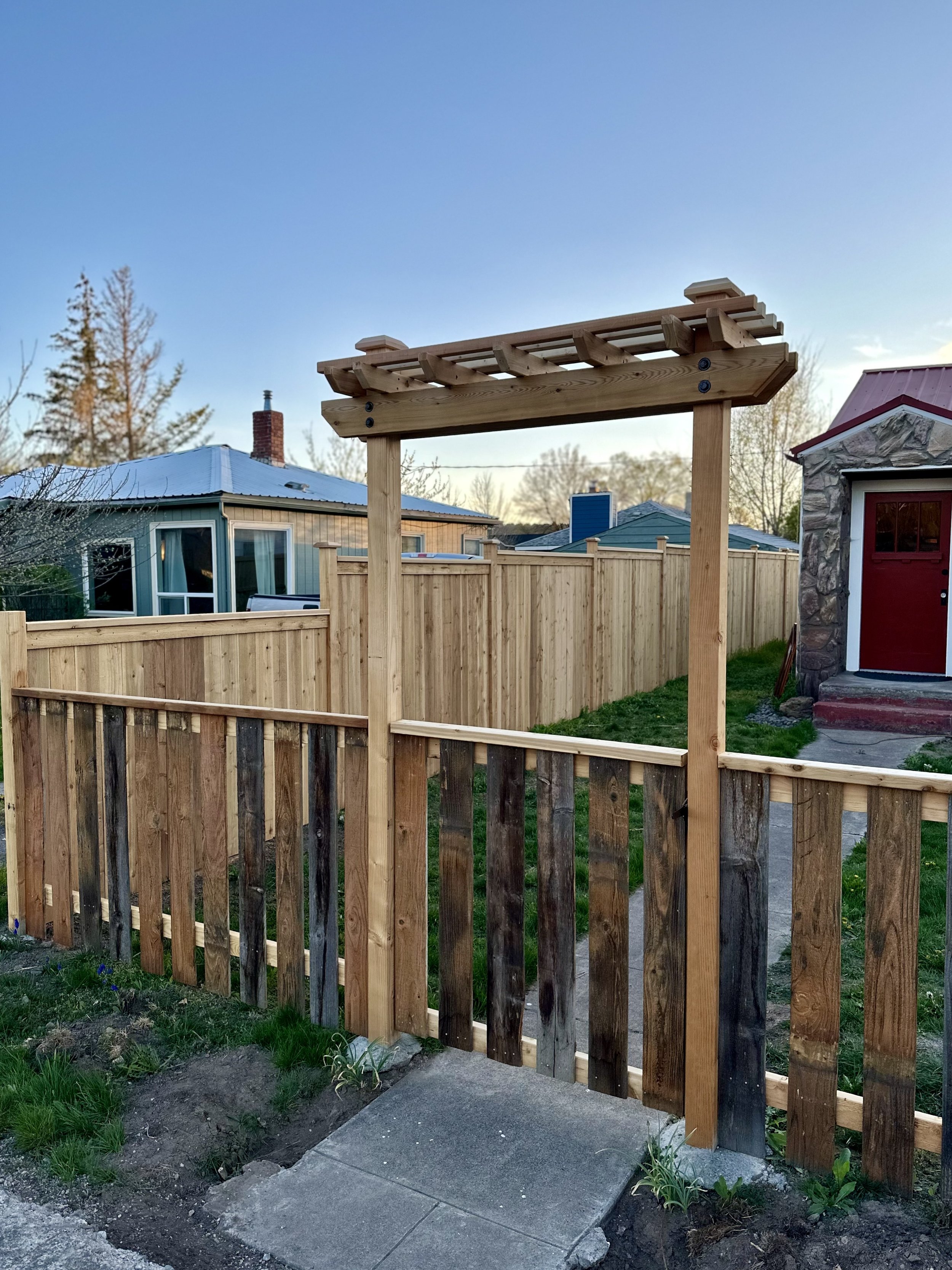 A wooden garden arbor frame in a backyard, surrounded by a partially built fence, with houses and trees in the background under a clear sky.