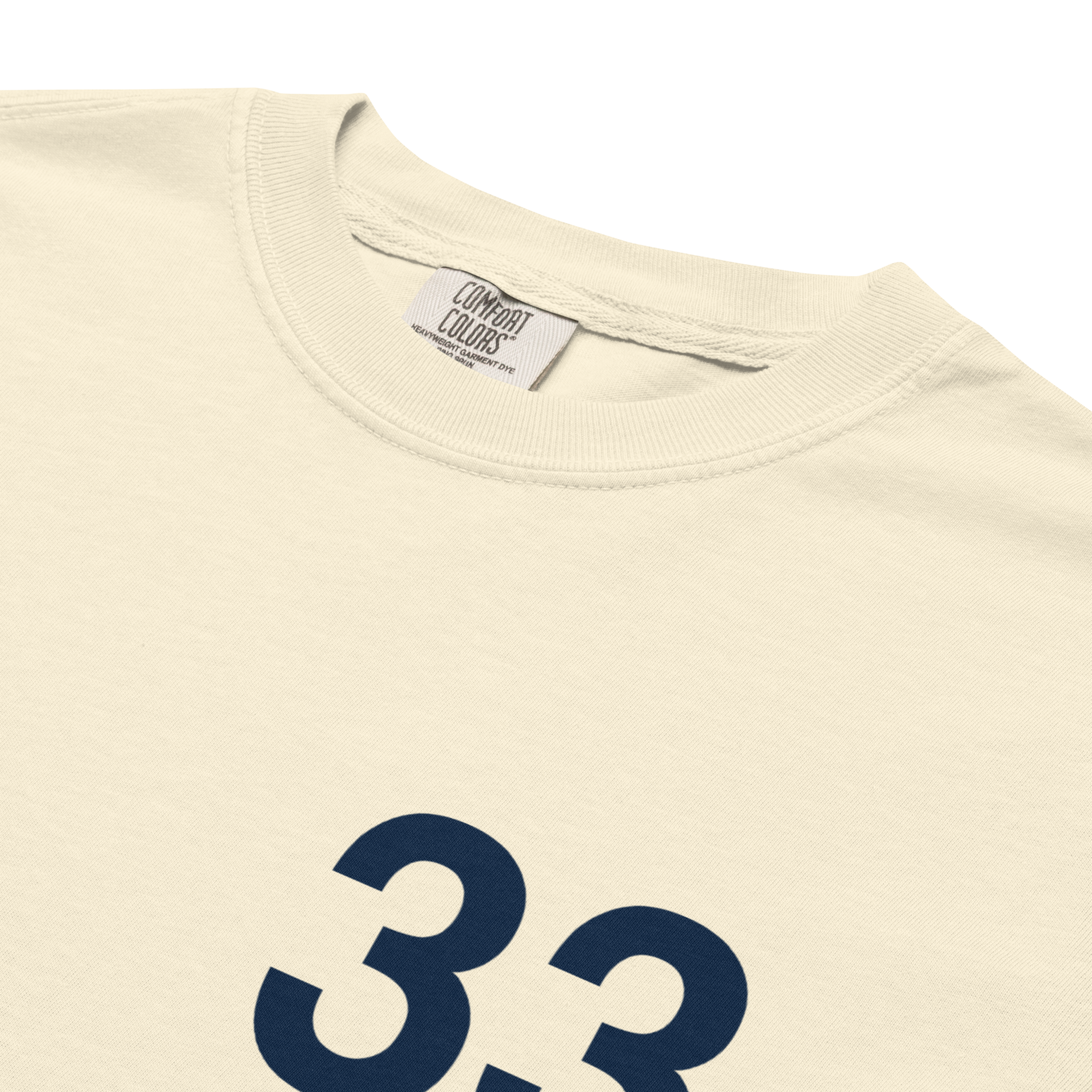 Close-up of the "33 & 7" chest print on a cream Comfort Colors heavyweight tee, showing the navy block numerals and the orange "the Mecca" handwritten script.