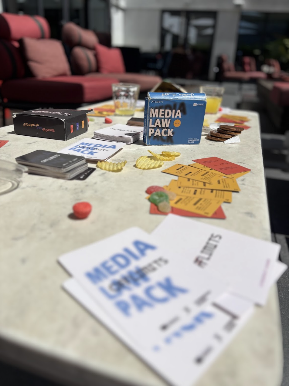 Board game setup on a table with cards, snacks, and a box labeled 'Media Law Pack'. There are red and yellow cards, some candies, and glasses of beverages, with outdoor seating in the background.