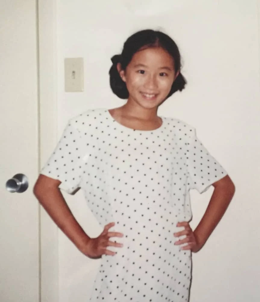 Yunzhe Zhou as a young girl with pigtails, shortly after immigrating to the US, wearing a white dress with small black polka dots, smiling with hands on hips, standing indoors against a plain white wall.