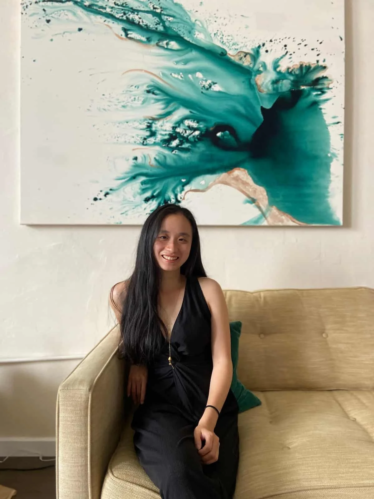 Yunzhe Zhou, business coach for Asian women, smiling while sitting on a sofa in front of an abstract teal and brown painting hanging on a white wall.