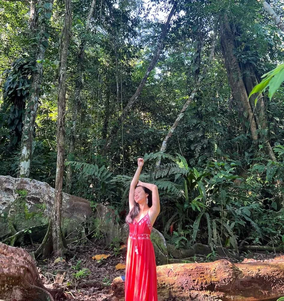 Yunzhe Zhou during her somatic facilitator training in Costa Rica, wearing a long red dress stands in a dense jungle, with her eyes closed and arms raised, one arm reaching upward and the other resting on her head, surrounded by lush green foliage.