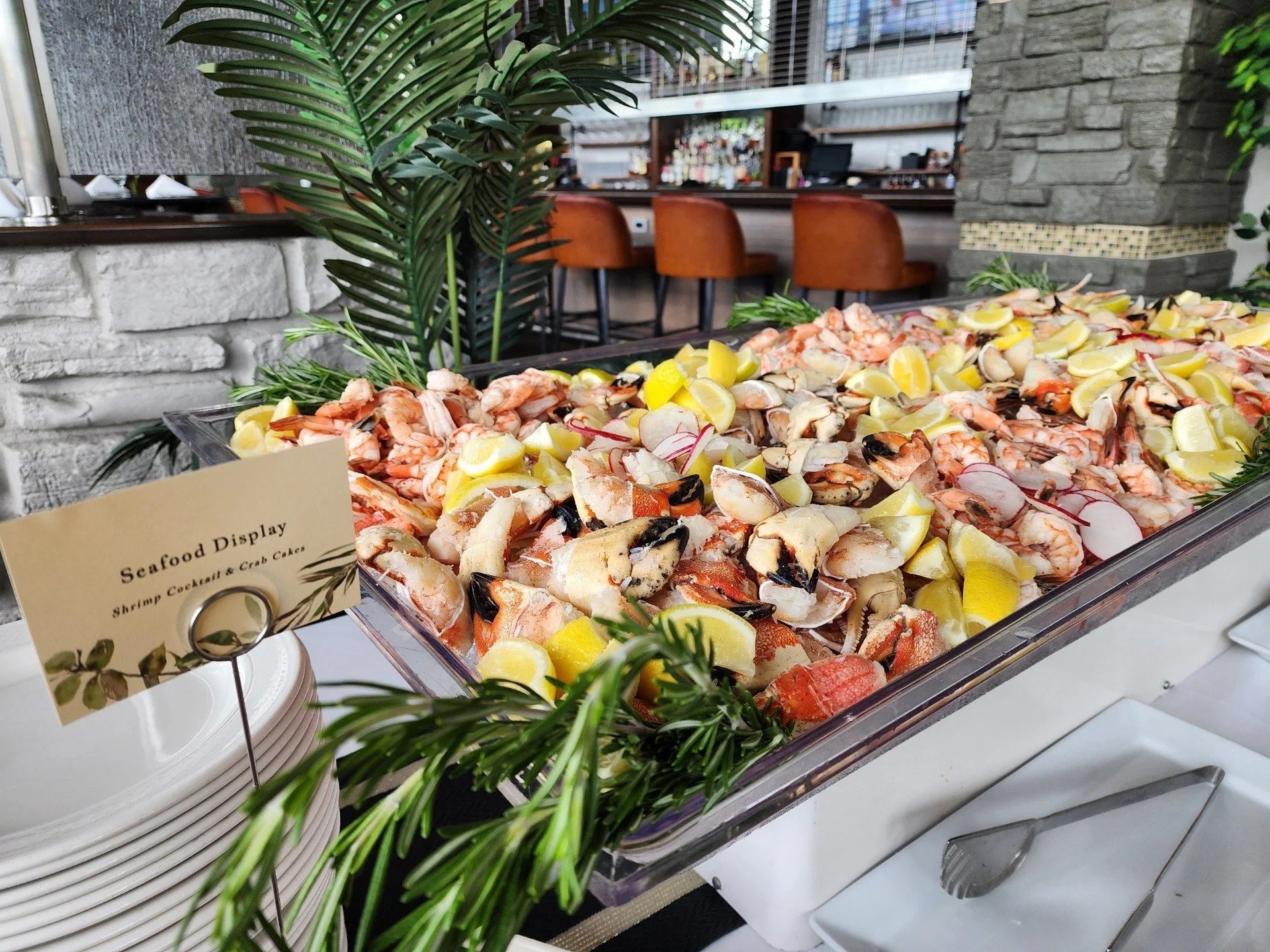 Seafood display with shrimp, crab cakes, lemon wedges, and radish slices at a buffet in a restaurant with bar chairs in the background.