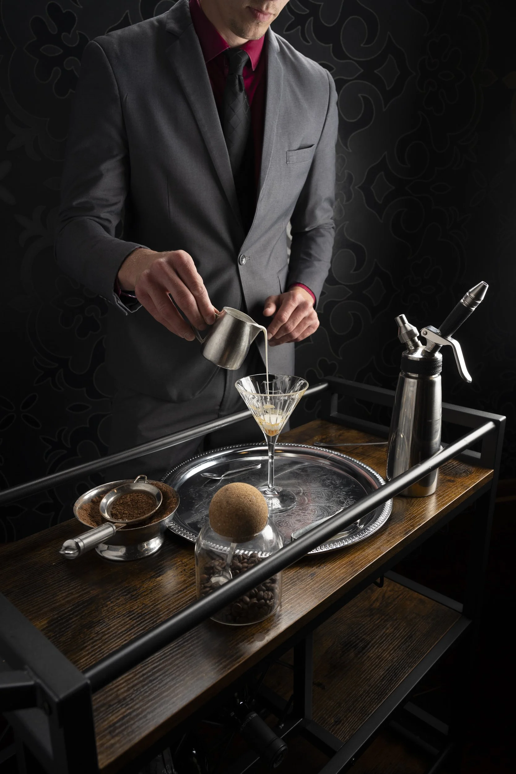 A man in a gray suit and red shirt pouring liquid into a cocktail glass on a wooden bar cart, with coffee beans, a strainer with ground coffee, a spray bottle, and an orange spherical object on the cart.