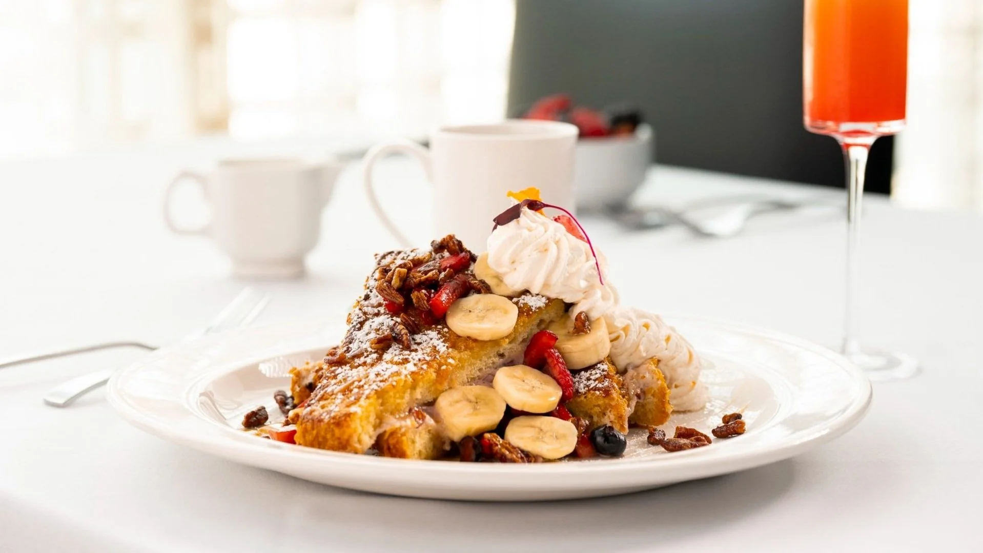 A slice of banana foster cake topped with whipped cream, banana slices, berries, nuts, and powdered sugar on a white plate, with coffee cups, a bowl of fruit, and a glass of red drink in the background on a white table.