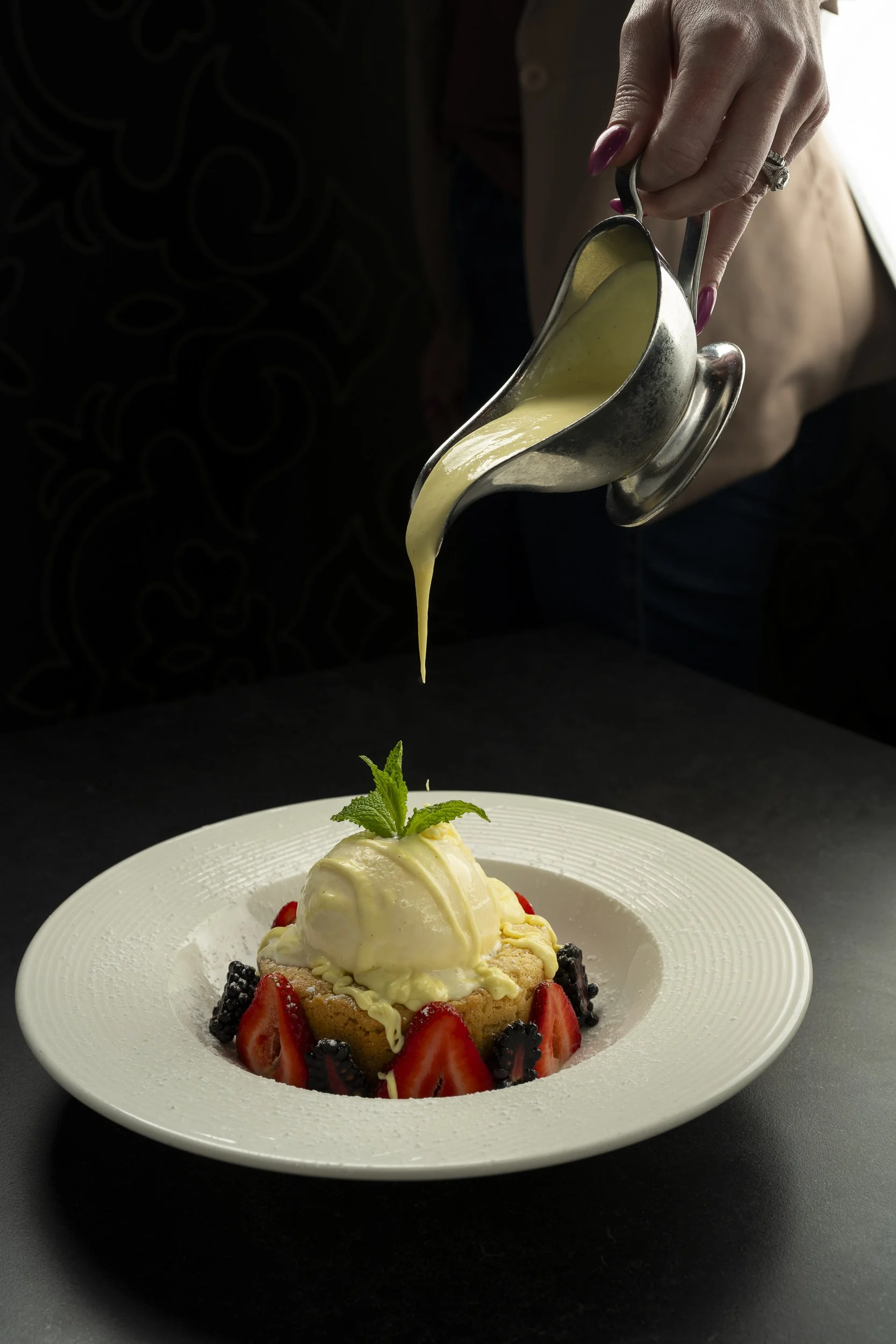 Person pouring vanilla sauce over a dessert topped with ice cream, strawberries, blackberries, and a mint leaf on a white plate.
