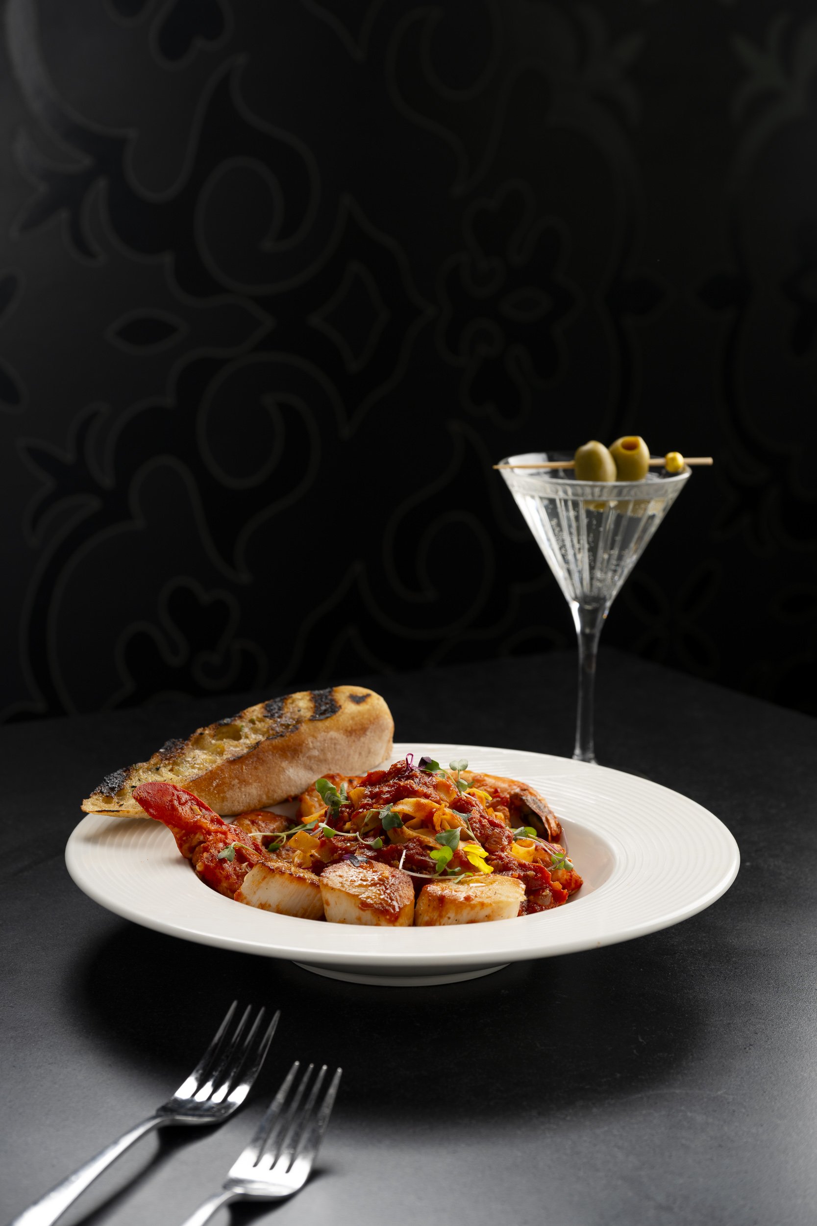 Plate of Italian food with gnocchi and grilled bread, black background with a martini glass holding green olives.