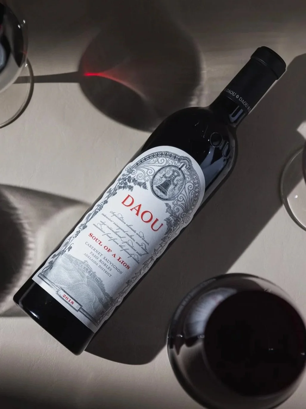 Two bottles of DAOU wine, one Cabernet Franc and one Cabernet Sauvignon, on a wooden surface with two glasses of red wine beside them in an outdoor setting with blurred background of trees and sunlight.