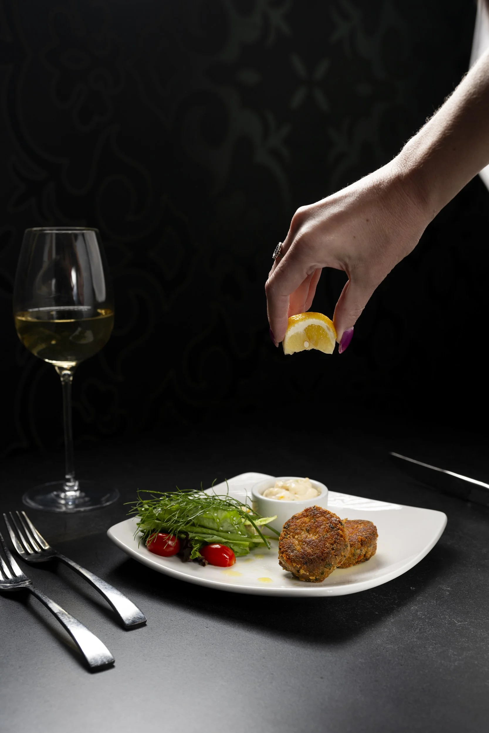 Hand squeezing lemon over a plate of food, which includes two croquettes, a small salad with tomatoes and greens, and a bowl of sauce, with a glass of white wine beside it.