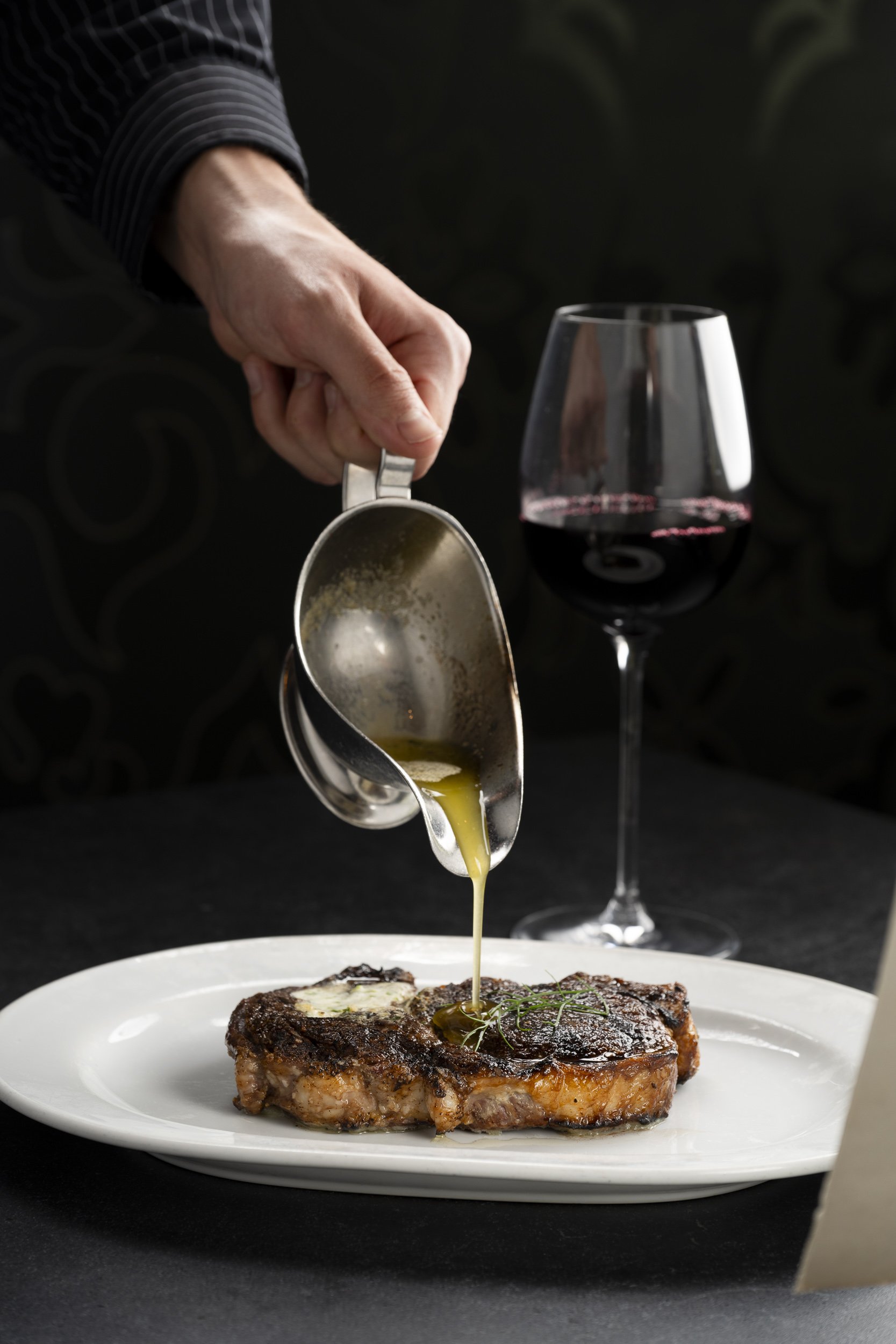 Person pouring sauce over a cooked steak on a white plate, with a glass of red wine in the background.