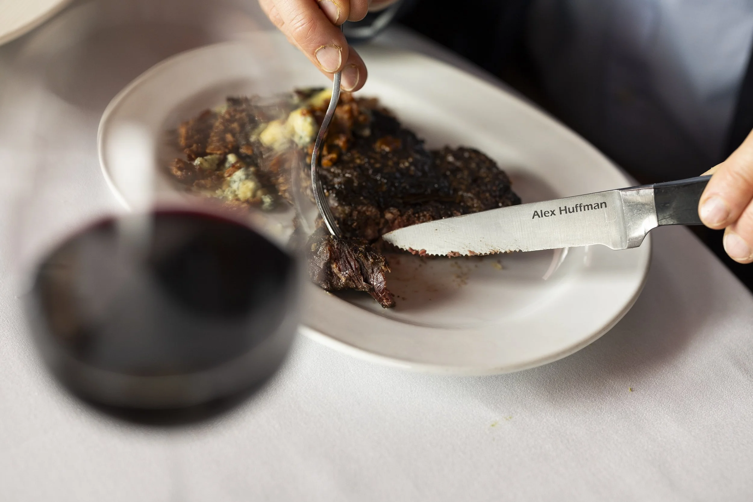 Person carving a cooked roast beef with a knife on a plate, with a glass of red wine in the foreground.
