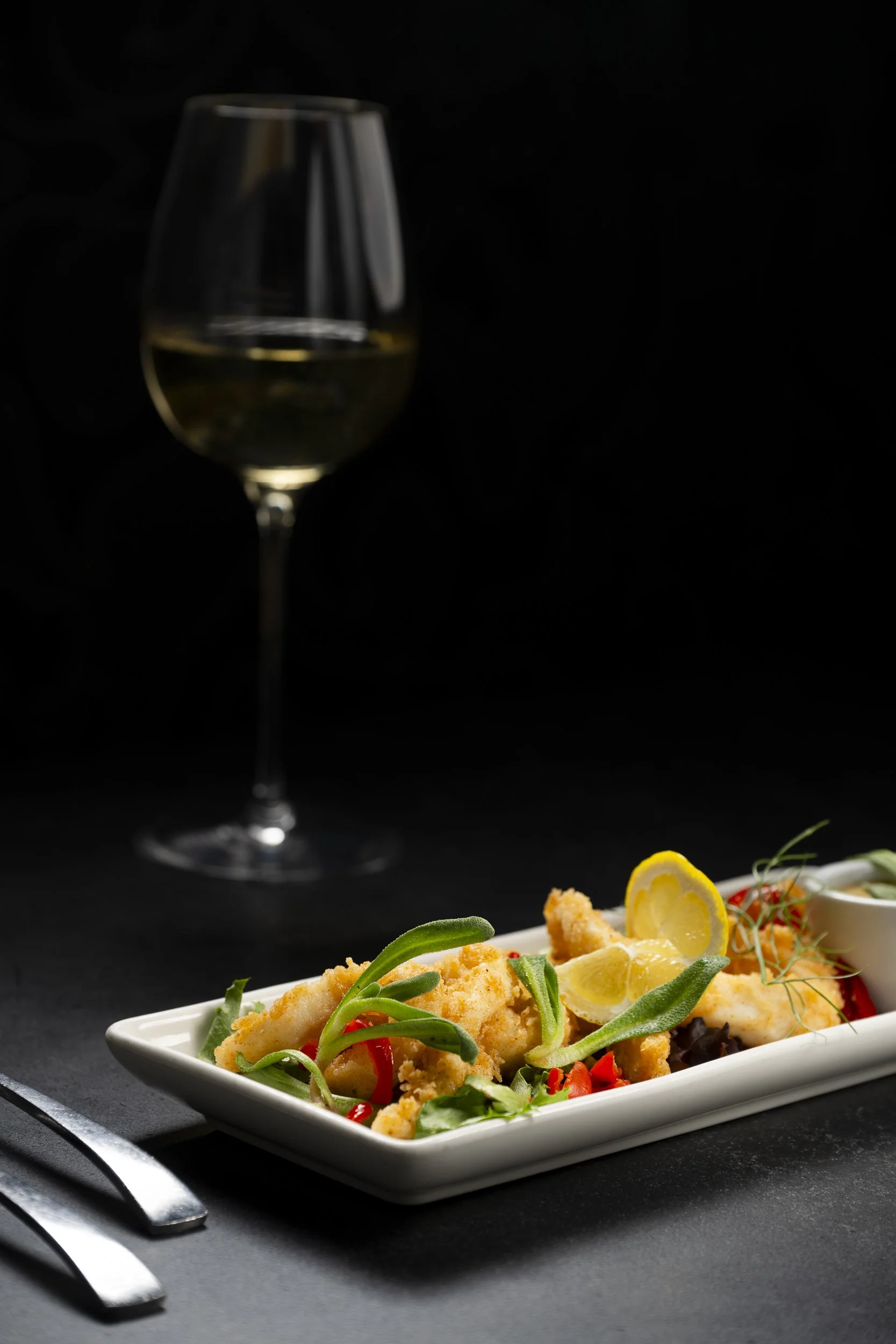 A plate of fried fish with lemon slices, garnished with greens, alongside a glass of white wine on a dark background.
