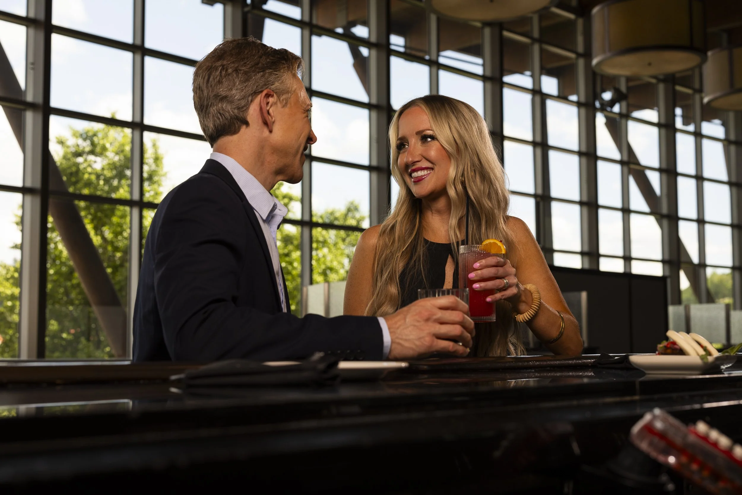 A man in a black suit and a woman with long blonde hair are smiling and talking at a bar, holding drinks. The woman has a glass with a pink beverage and an orange slice garnish.