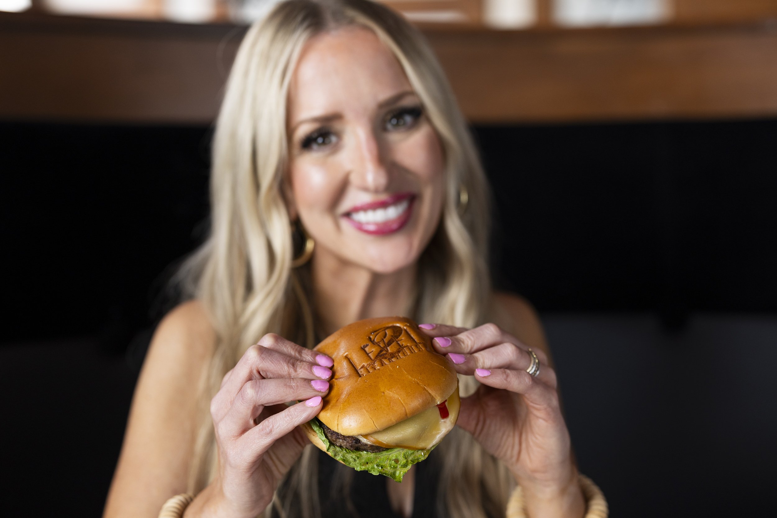 A woman with long blonde hair holding a cheeseburger with lettuce, cheese, and ketchup, smiling in a restaurant.