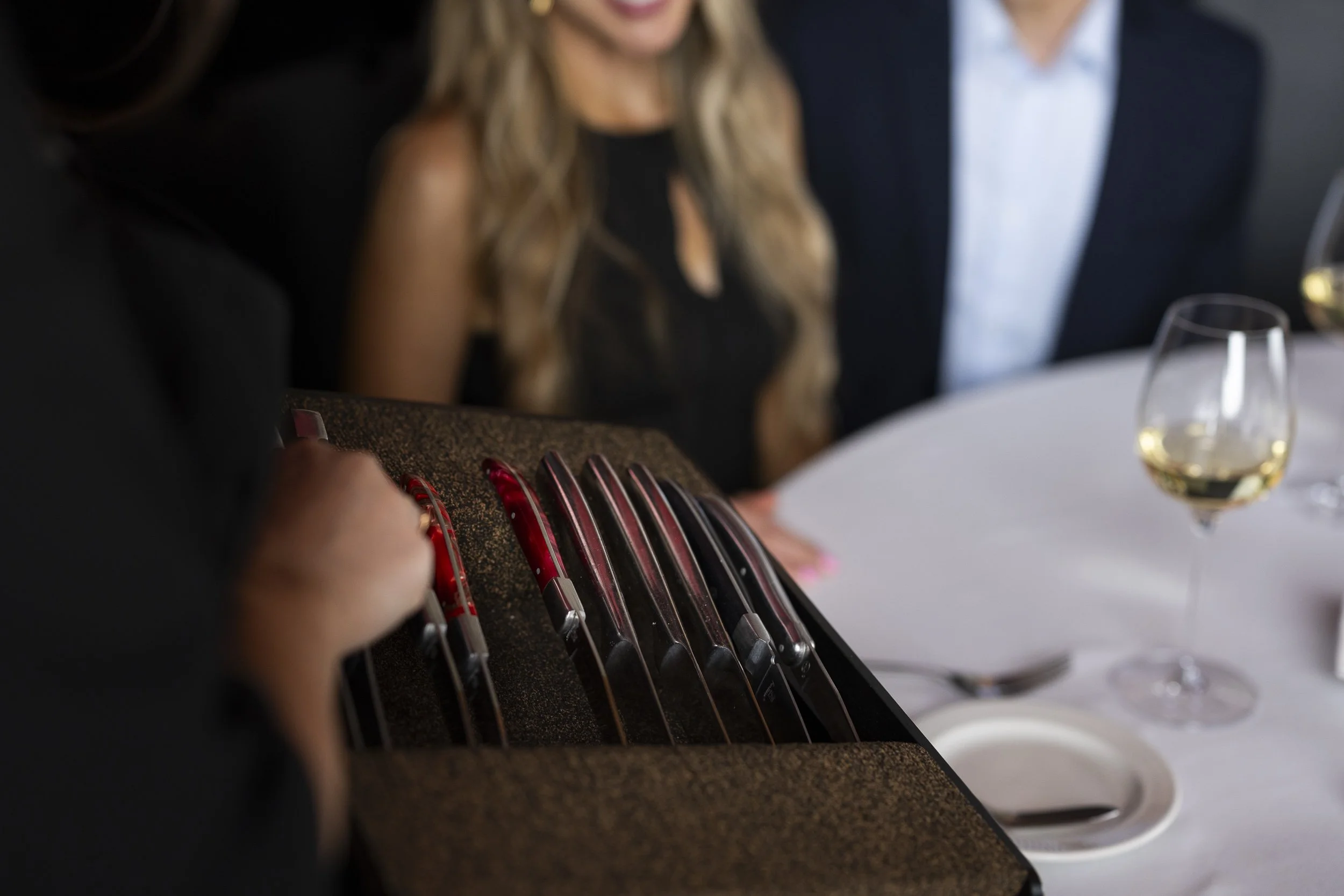 A person in black holding a tray of different colored knives, with a table set for a meal and a woman with long hair and a black dress in the background.