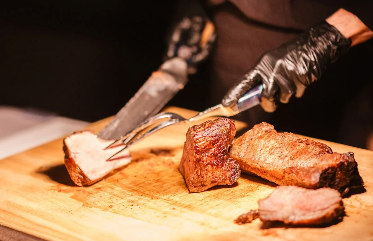 Person slicing cooked meat with a carving fork and knife on a wooden cutting board.