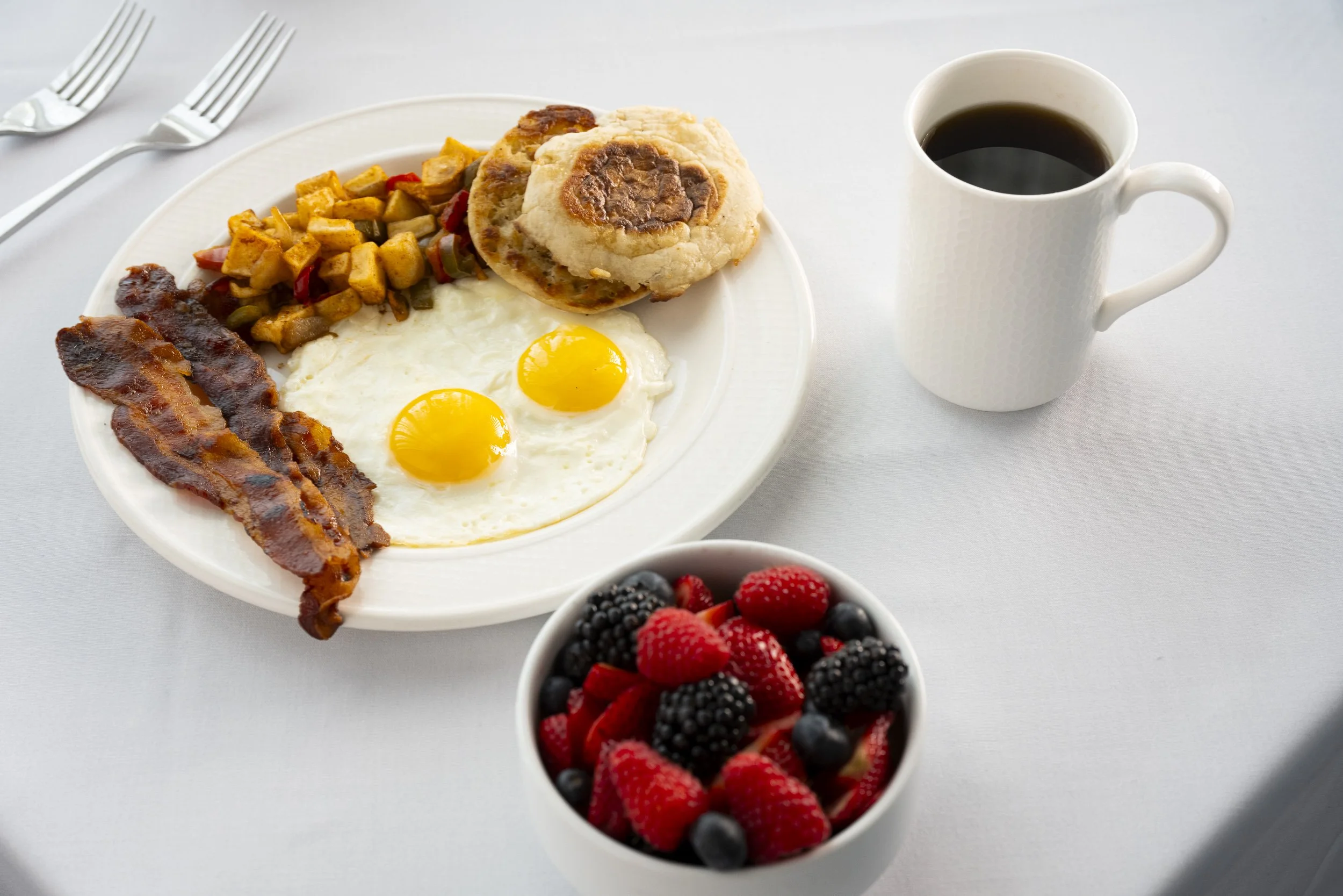 A breakfast plate with two fried eggs, bacon, breakfast sausage with biscuit, roasted potatoes, served with a mug of black coffee and a bowl of mixed berries on a white table with two forks.