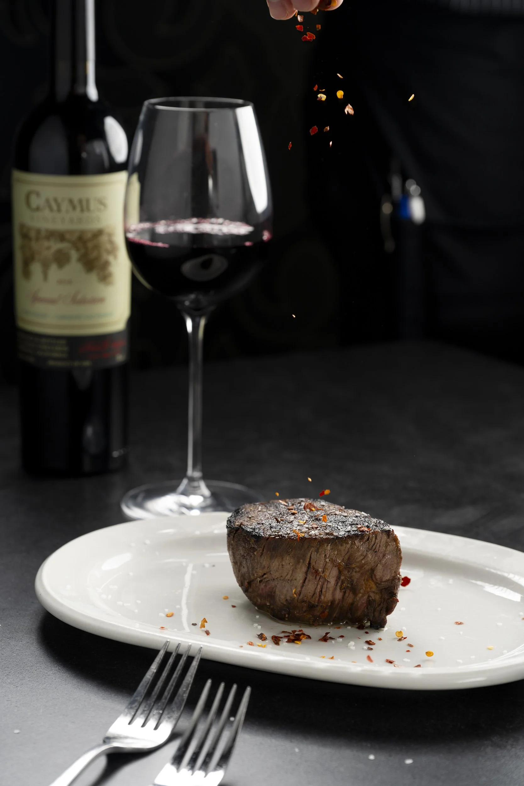 A cooked steak on a white plate, a glass of red wine, and a bottle of Caymus wine in the background.