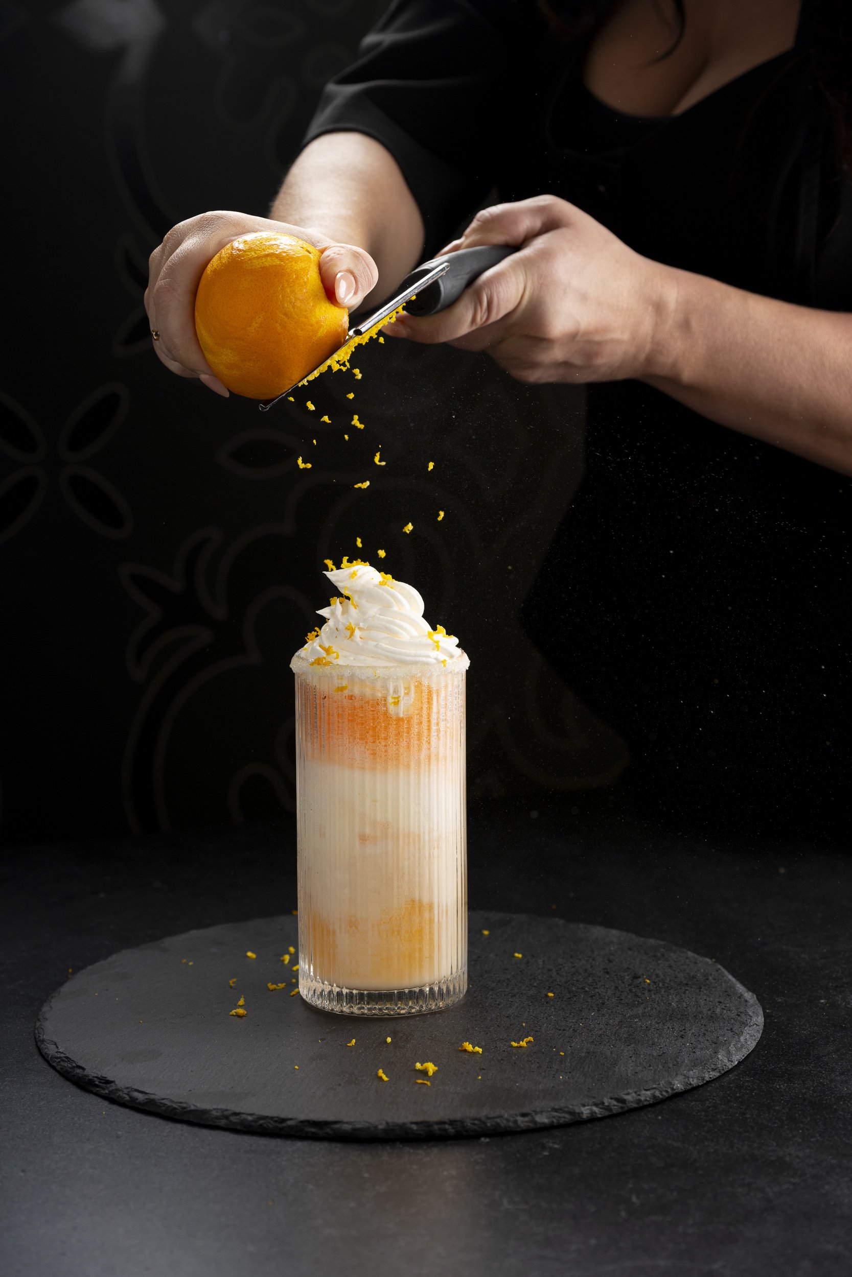A person grating an orange over a cream-colored beverage topped with whipped cream in a tall glass with an orange tint on a black slate coaster.