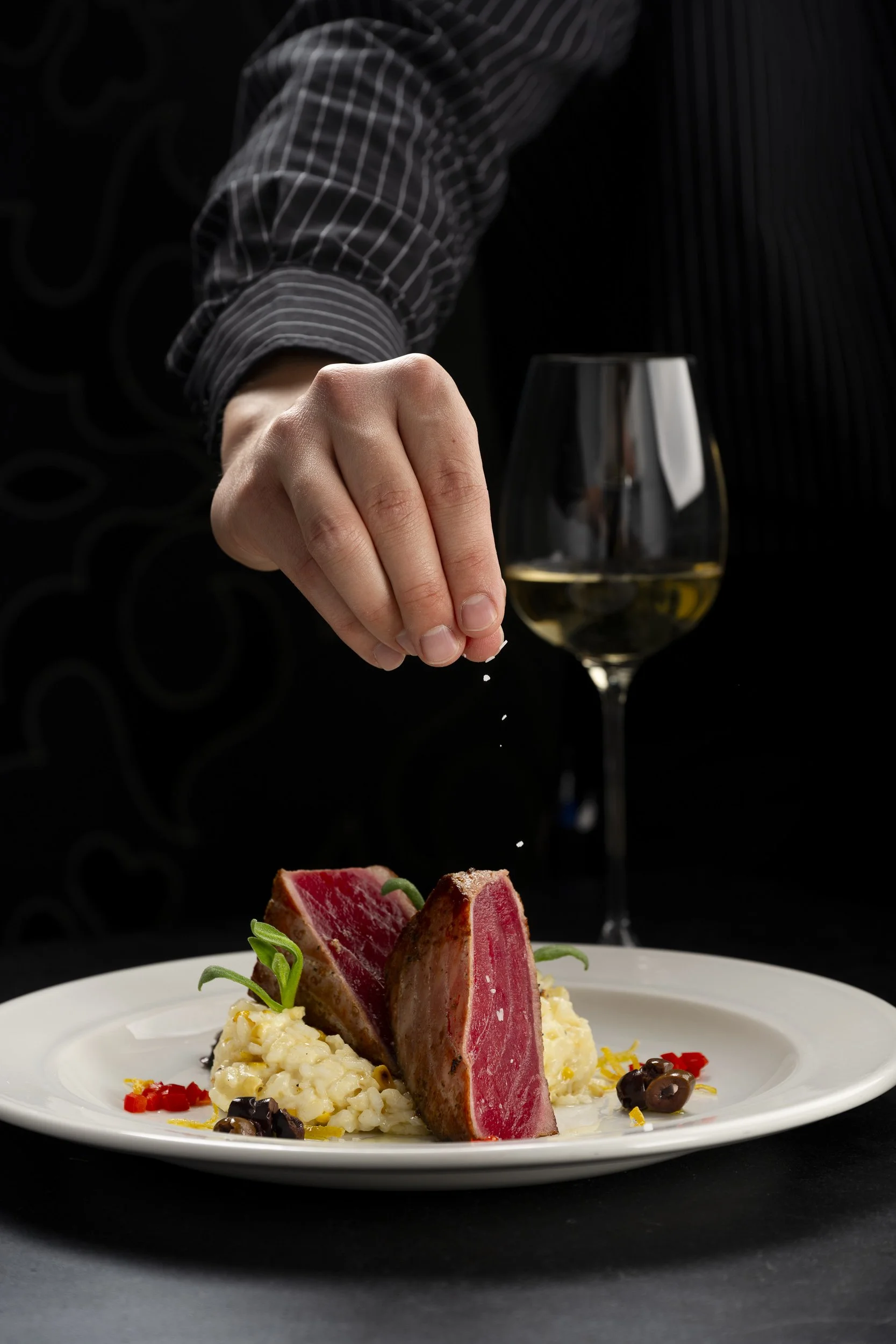 A person sprinkles salt on a gourmet dish of seared tuna steak served with mashed potatoes, garnished with greens and olives, with a glass of white wine in the background.