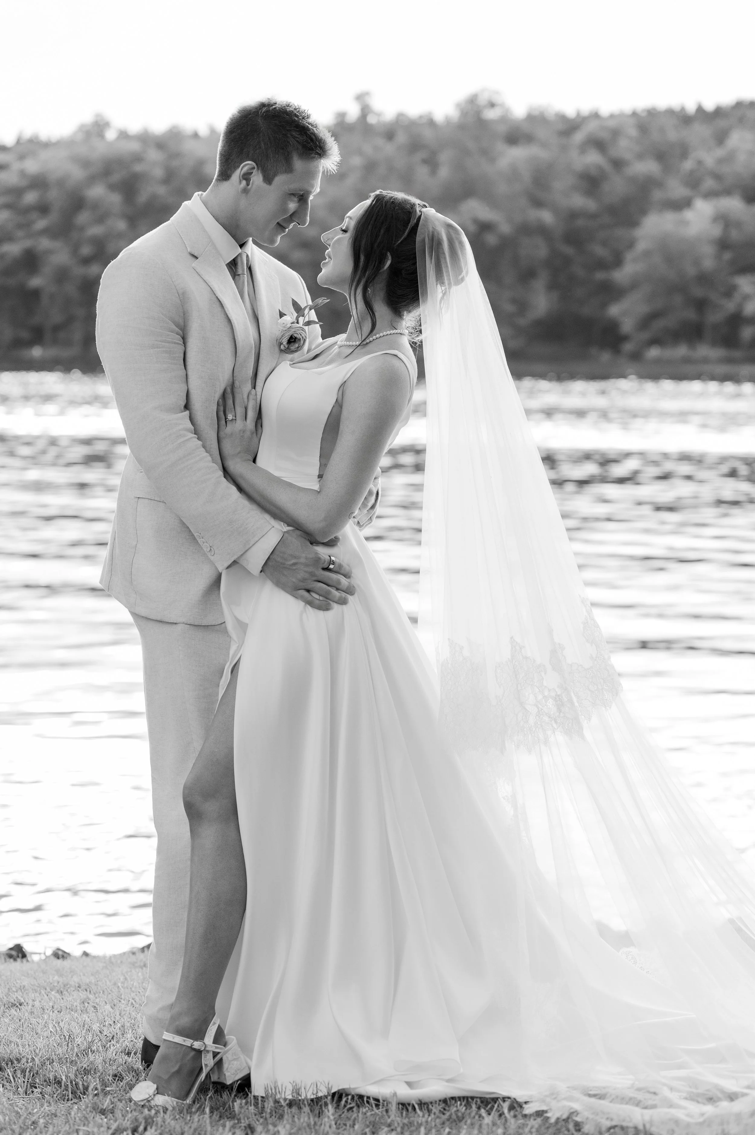 A black and white photograph of a bride and groom standing close together outdoors near a body of water, looking into each other's eyes, with a blurred tree-lined background.