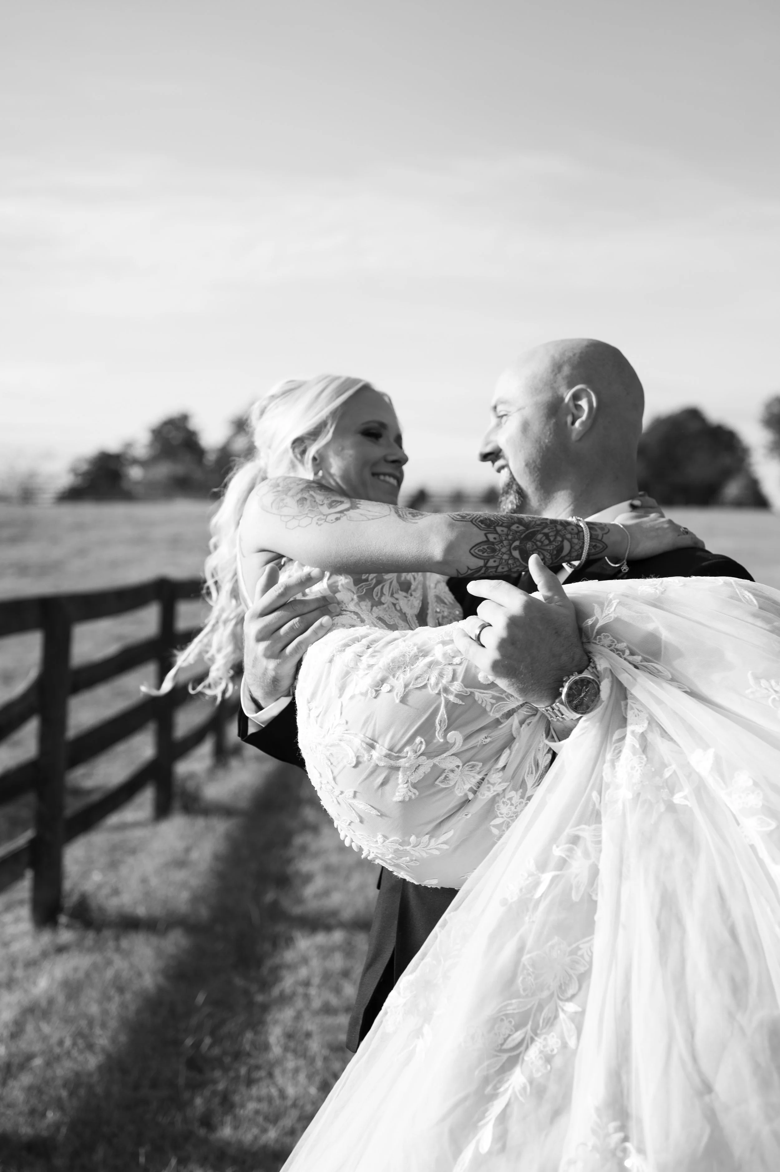 A couple, possibly newlyweds, sharing a joyful moment outdoors, with the man lifting the woman in his arms. The woman is wearing a wedding dress and has tattoos on her arms. The background is a broad landscape with trees and a fence.