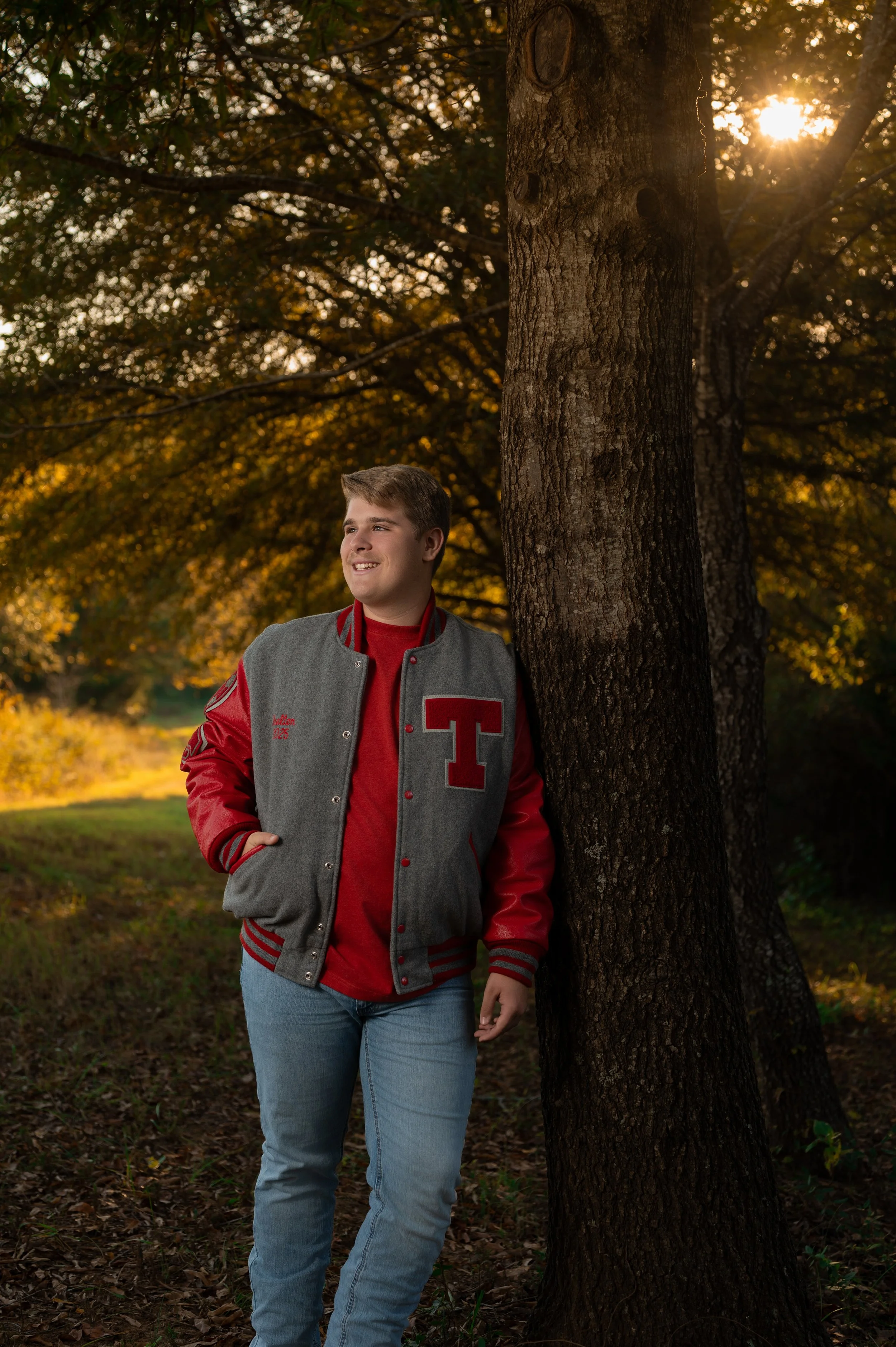A young man wearing a gray and red letterman jacket and light blue jeans stands next to a tree in a park during autumn, with sunlight filtering through the leaves.