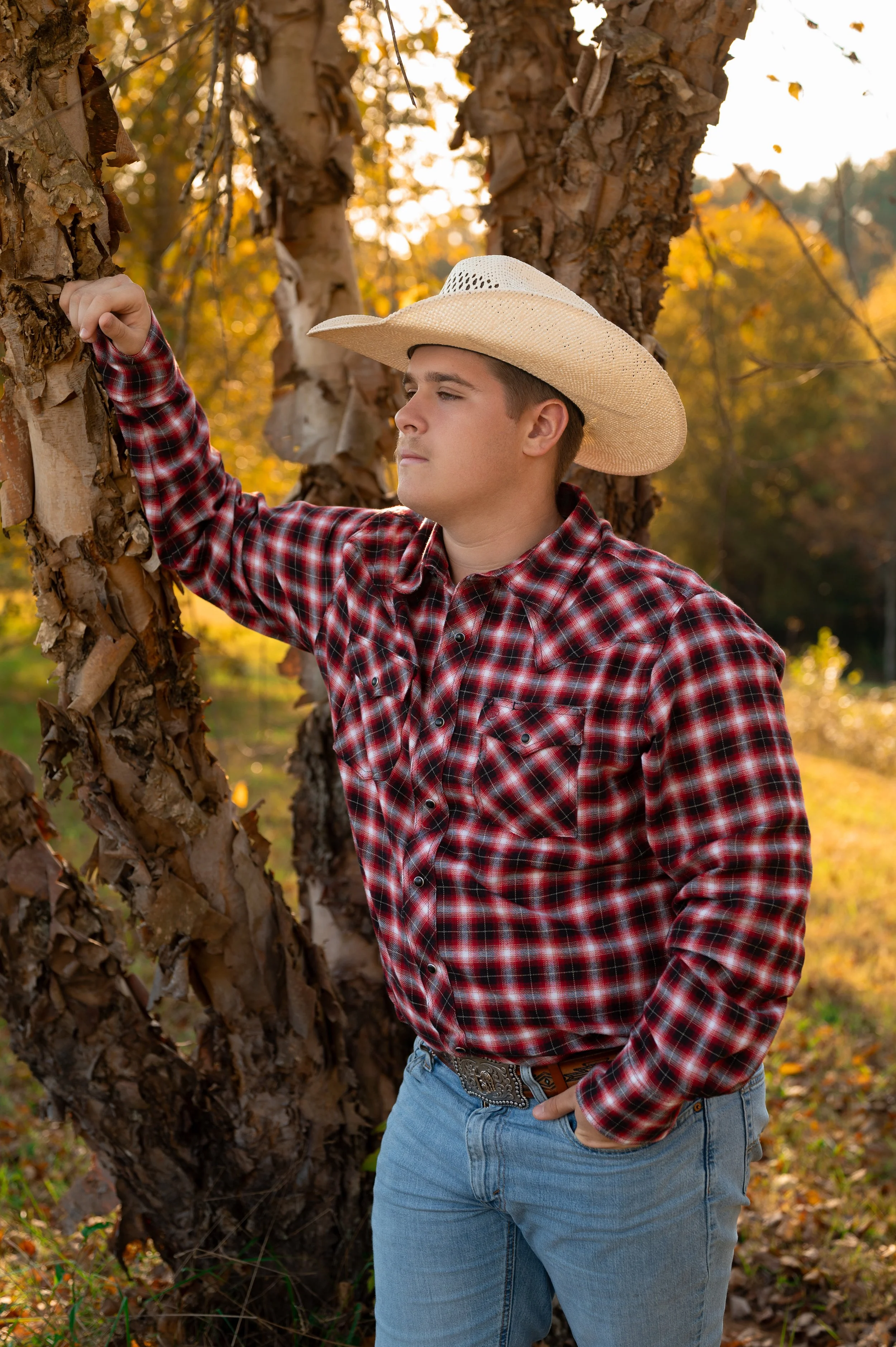 A young man wearing a plaid shirt and cowboy hat stands outdoors in front of a tree, looking contemplative.