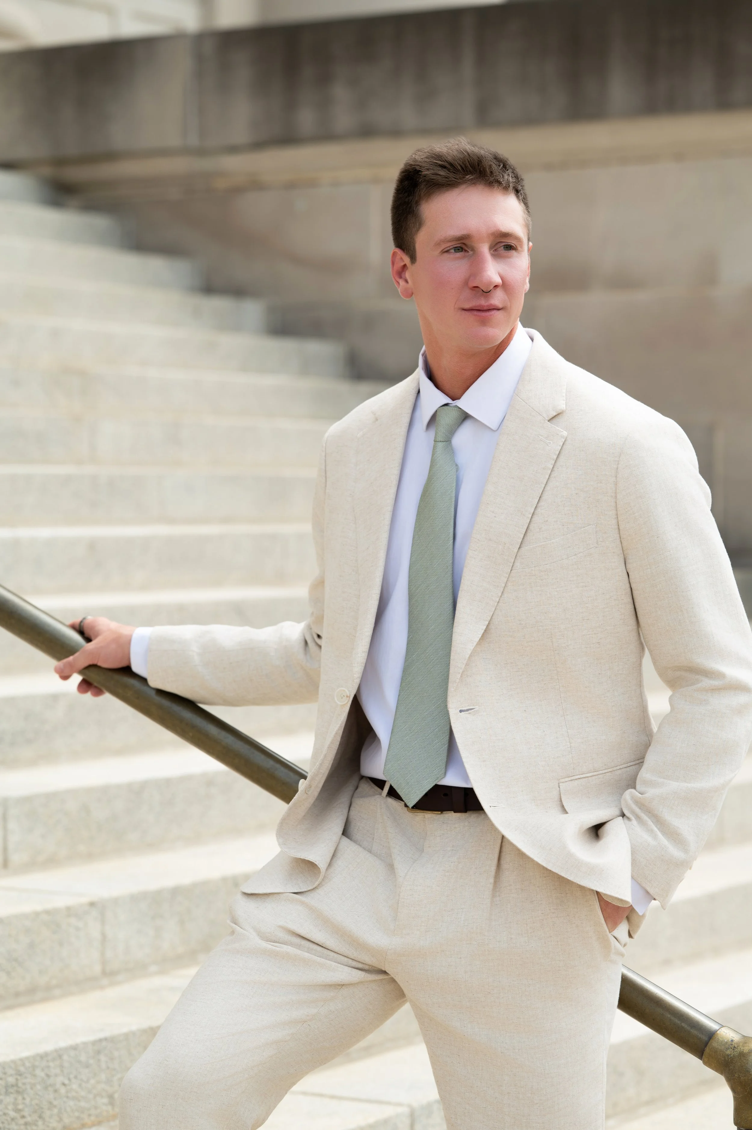 A young man in a light-colored suit and tie standing outside on stairs, leaning on a handrail, with one hand in his pocket, looking to the side.