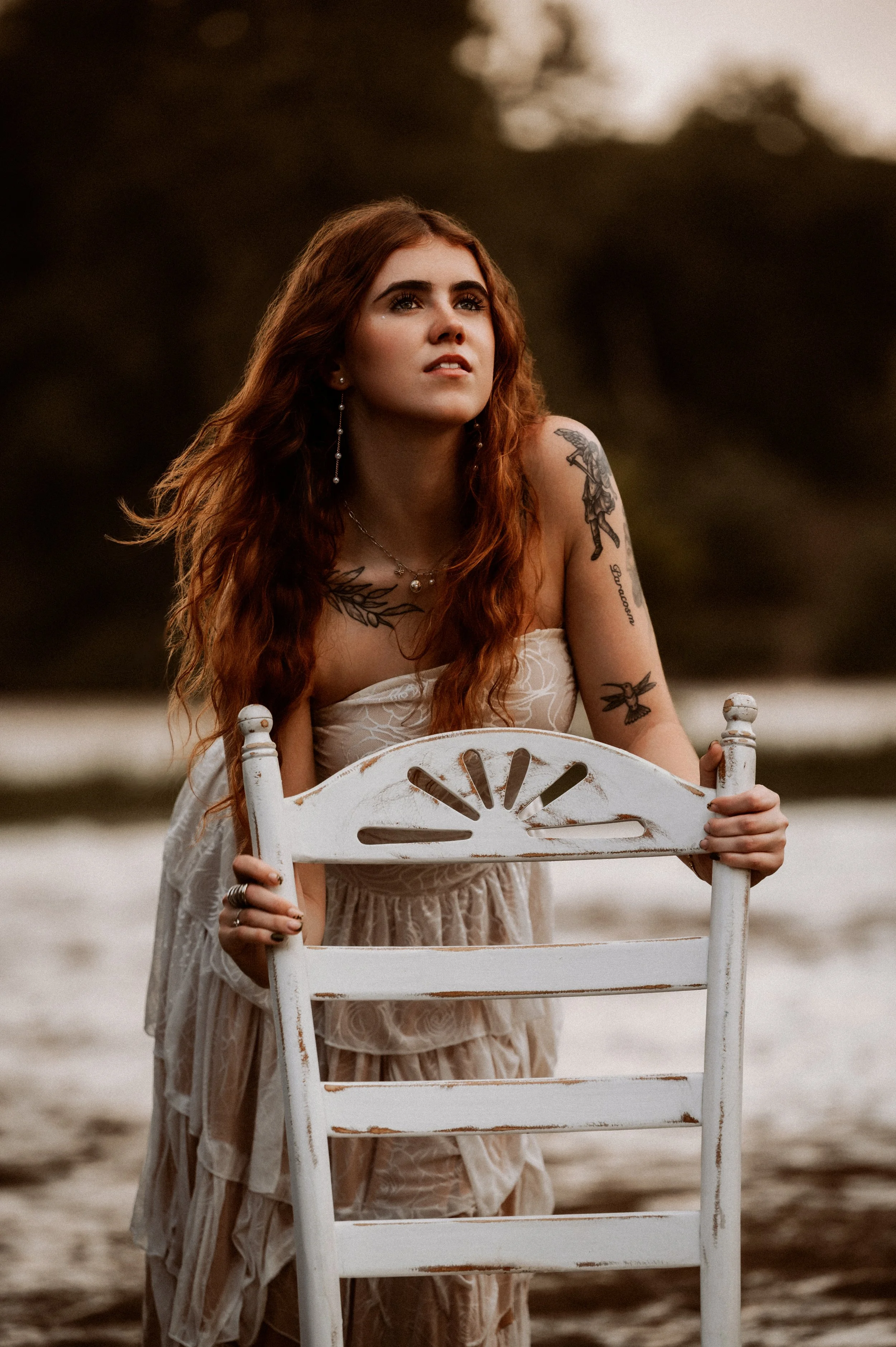 A young woman with long, wavy red hair and tattoos on her arms, wearing a strapless dress, holding a white wooden chair on a beach at sunset, looking into the distance.