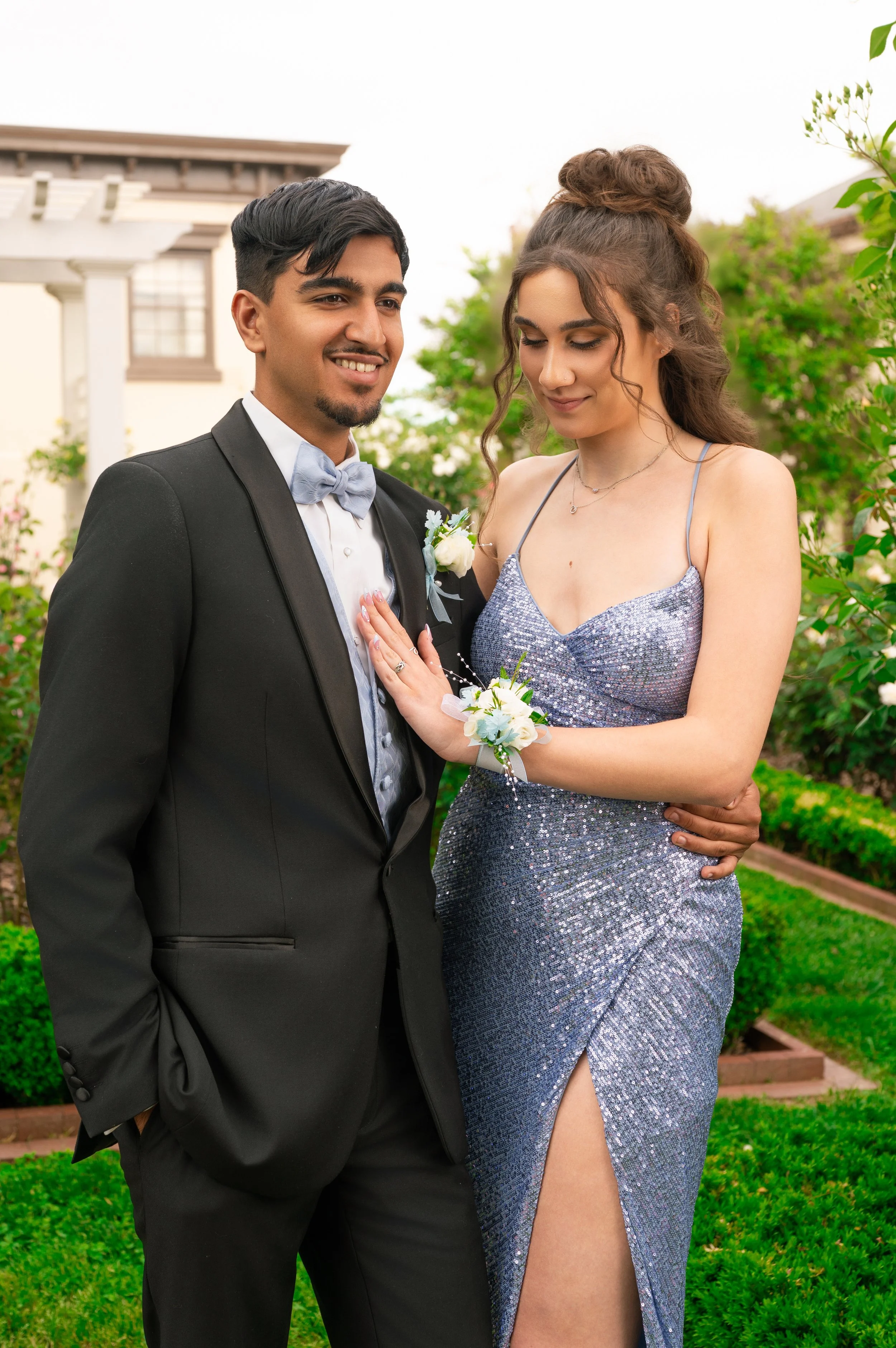 A young man in a black tuxedo with a light blue bow tie and boutonniere stands smiling next to a young woman in a sparkly silver evening gown with a thigh-high slit, corsage, and a bun hairstyle, outdoors on a garden lawn.