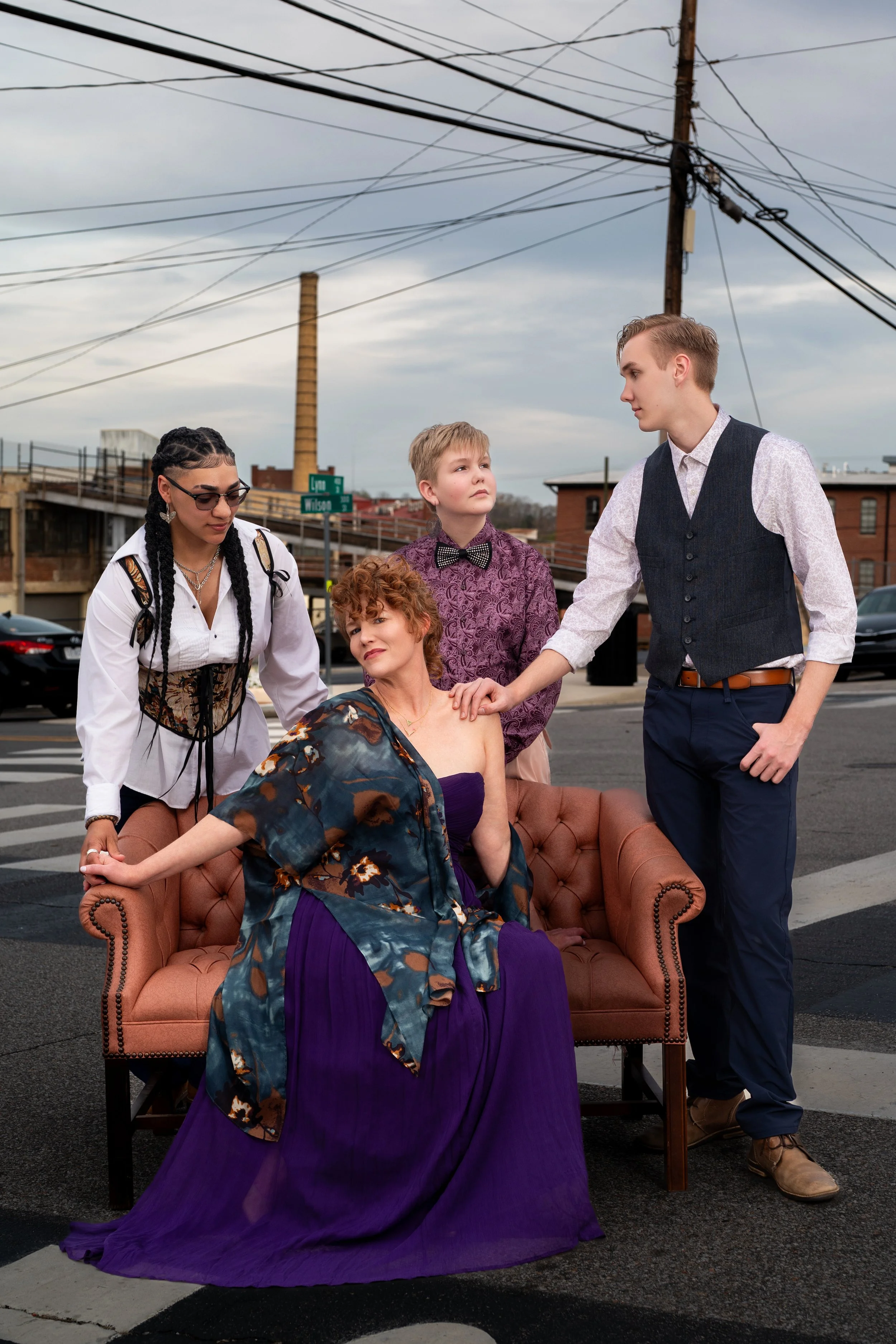 Four people on a street, with an urban background, gathered around a woman sitting on a vintage sofa. The woman wears a purple dress and a patterned shawl, looking thoughtful. A young man in a vest and white shirt touches her shoulder, while a boy wi