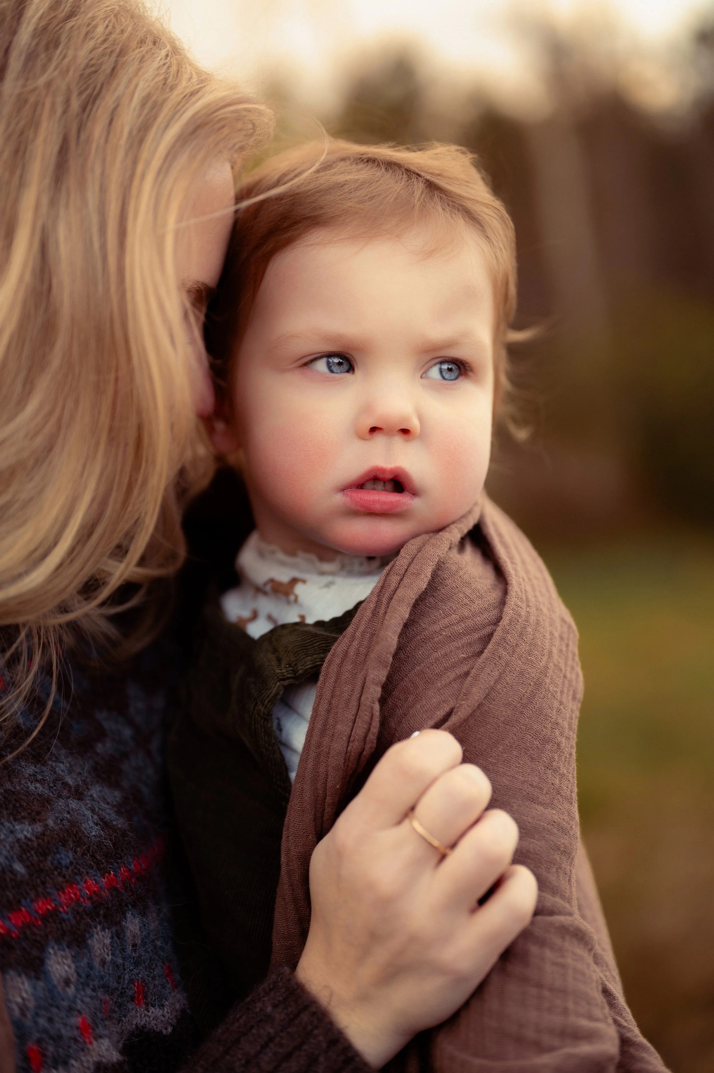 A woman with blonde hair holding a young boy with blonde hair and blue eyes outdoors, during sunset or late afternoon, with a blurred natural background.
